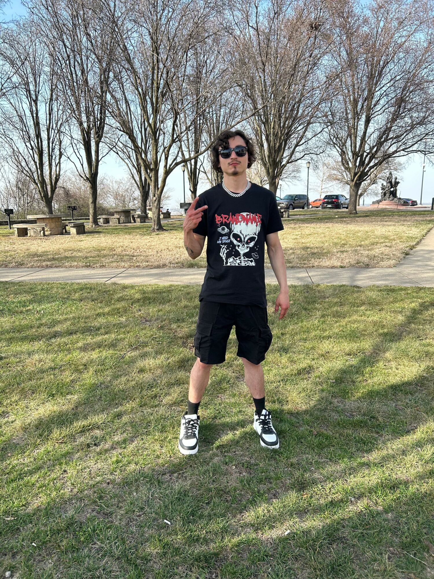 Young person standing on grass in a park, wearing sunglasses, black t-shirt, shorts, and sneakers, with trees in the background.