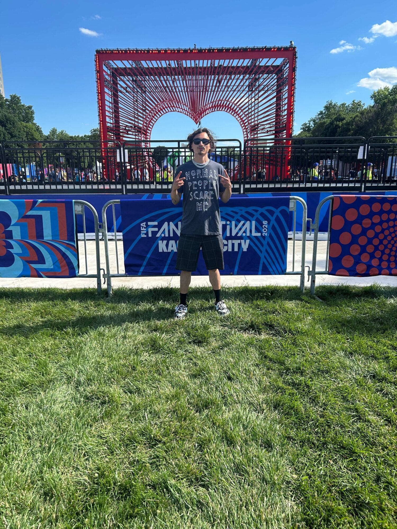 Person standing in front of a colorful heart-shaped structure at an outdoor festival, with trees and blue sky in background.