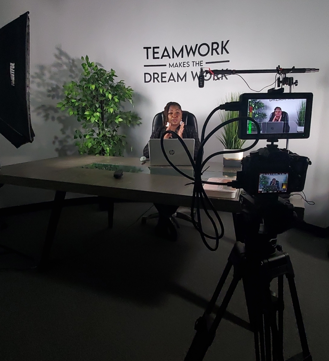 Person sitting at a desk with a camera and monitor recording, wall with motivational quote, and a plant in the background.