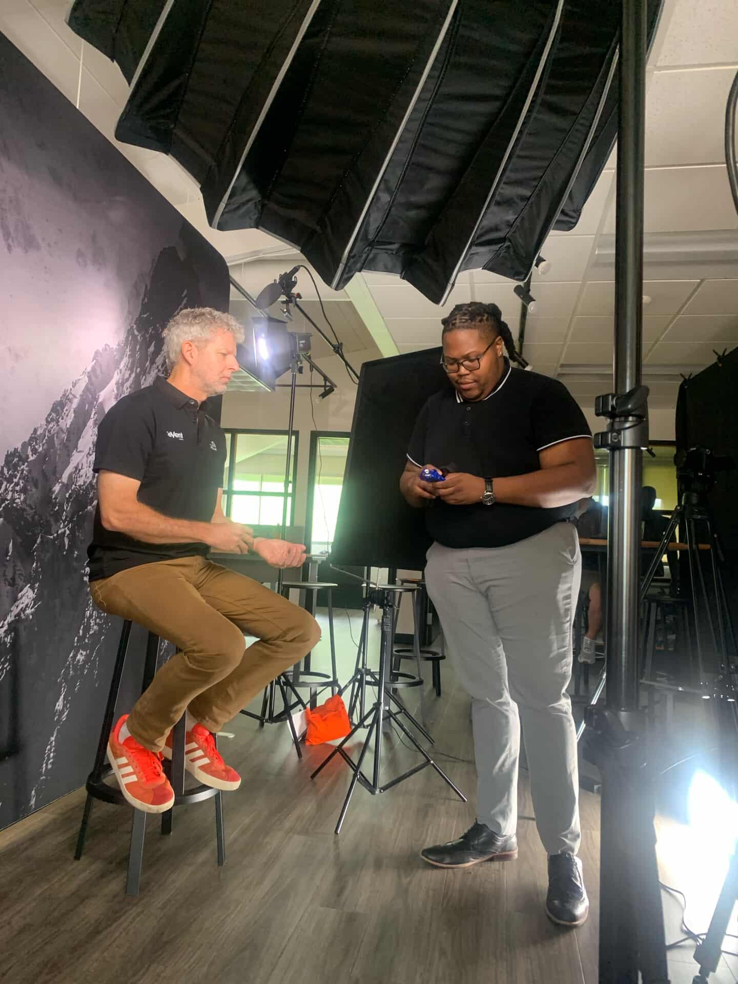 Two men are in a studio with lighting equipment, one seated on a stool and the other standing, both looking at a phone.