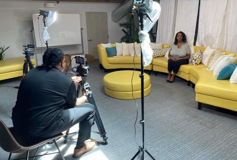 Person being filmed sitting on a yellow sofa in a studio with filming equipment and a camera operator.