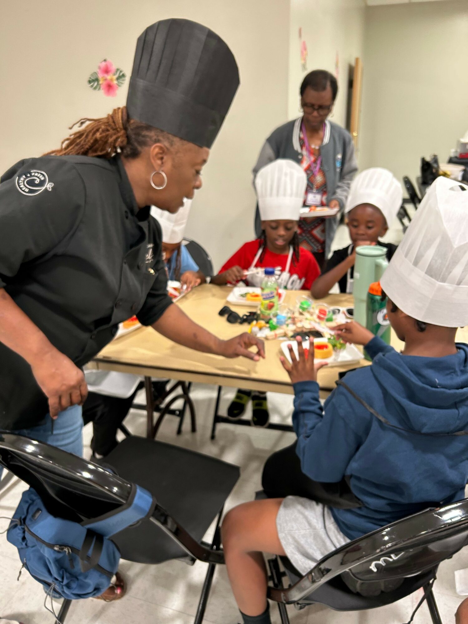 Person wearing a chef's hat and black shirt reaching for a birthday cake with colorful decorations, surrounded by children and adults.