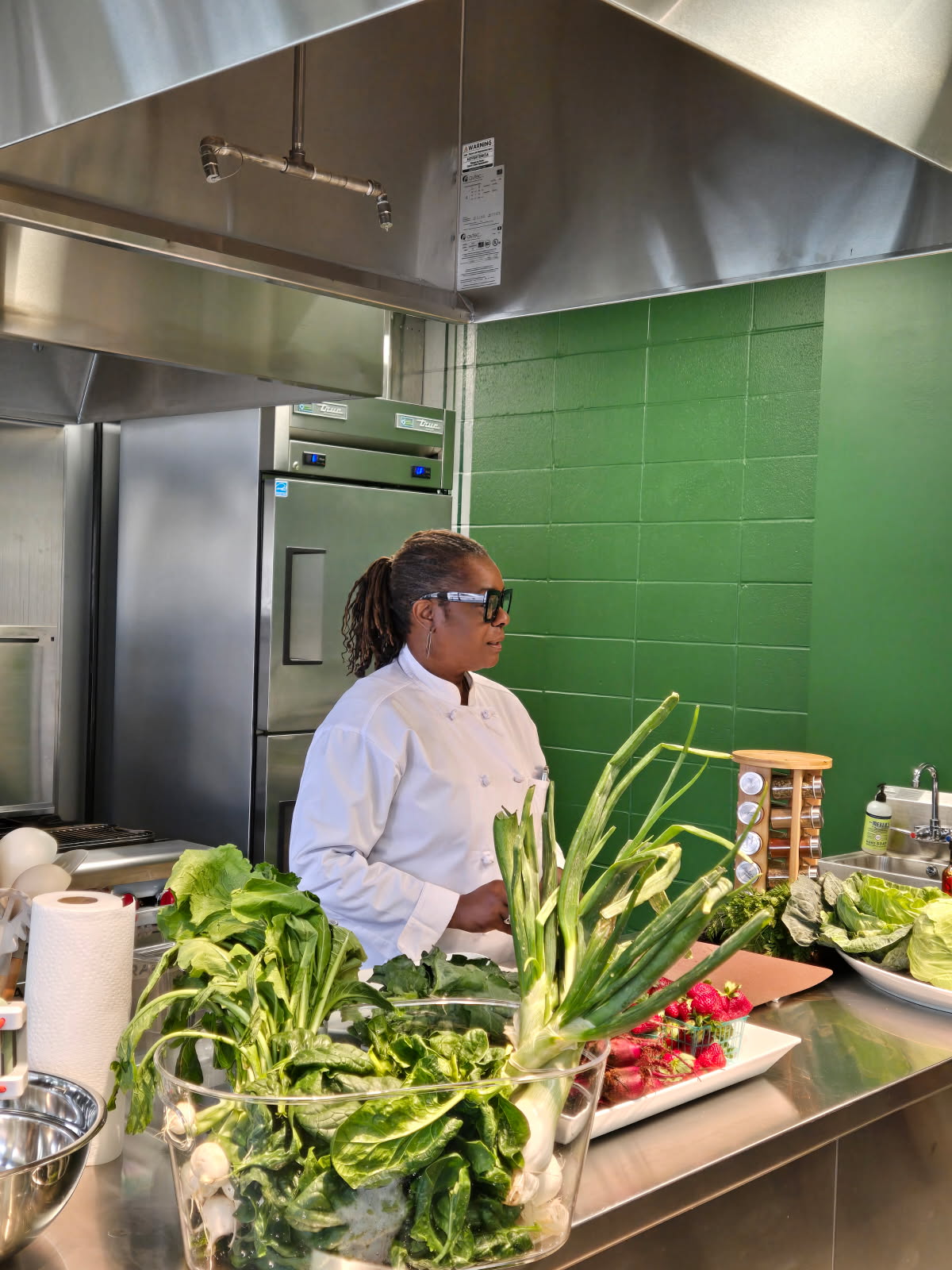 A woman in a white chef's coat and sunglasses stands in a kitchen with green tiled wall, surrounded by vegetables and kitchen tools.