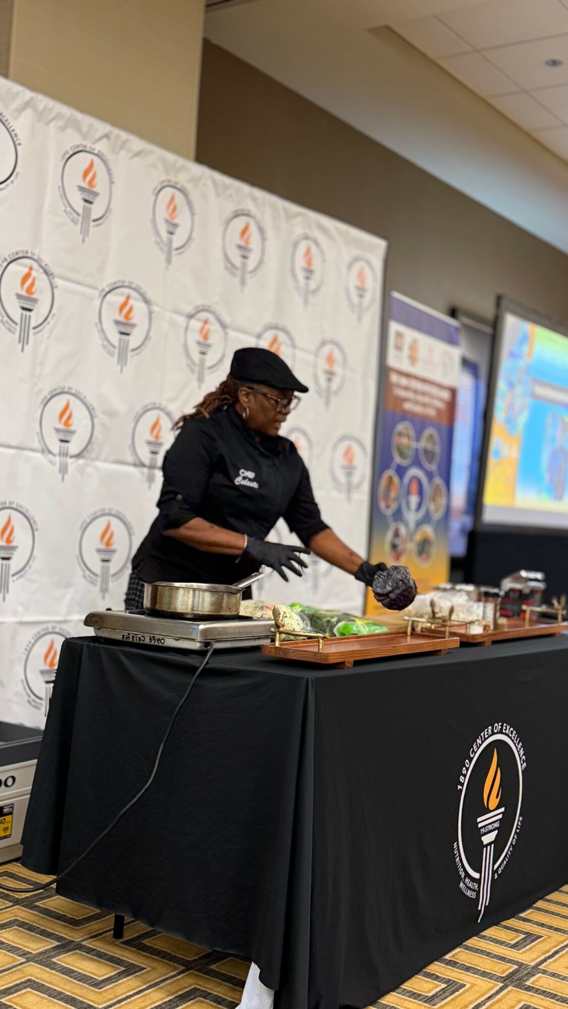 Person wearing black apron and hat preparing food at a table with a black tablecloth, backdrop with logo, and screens in background.