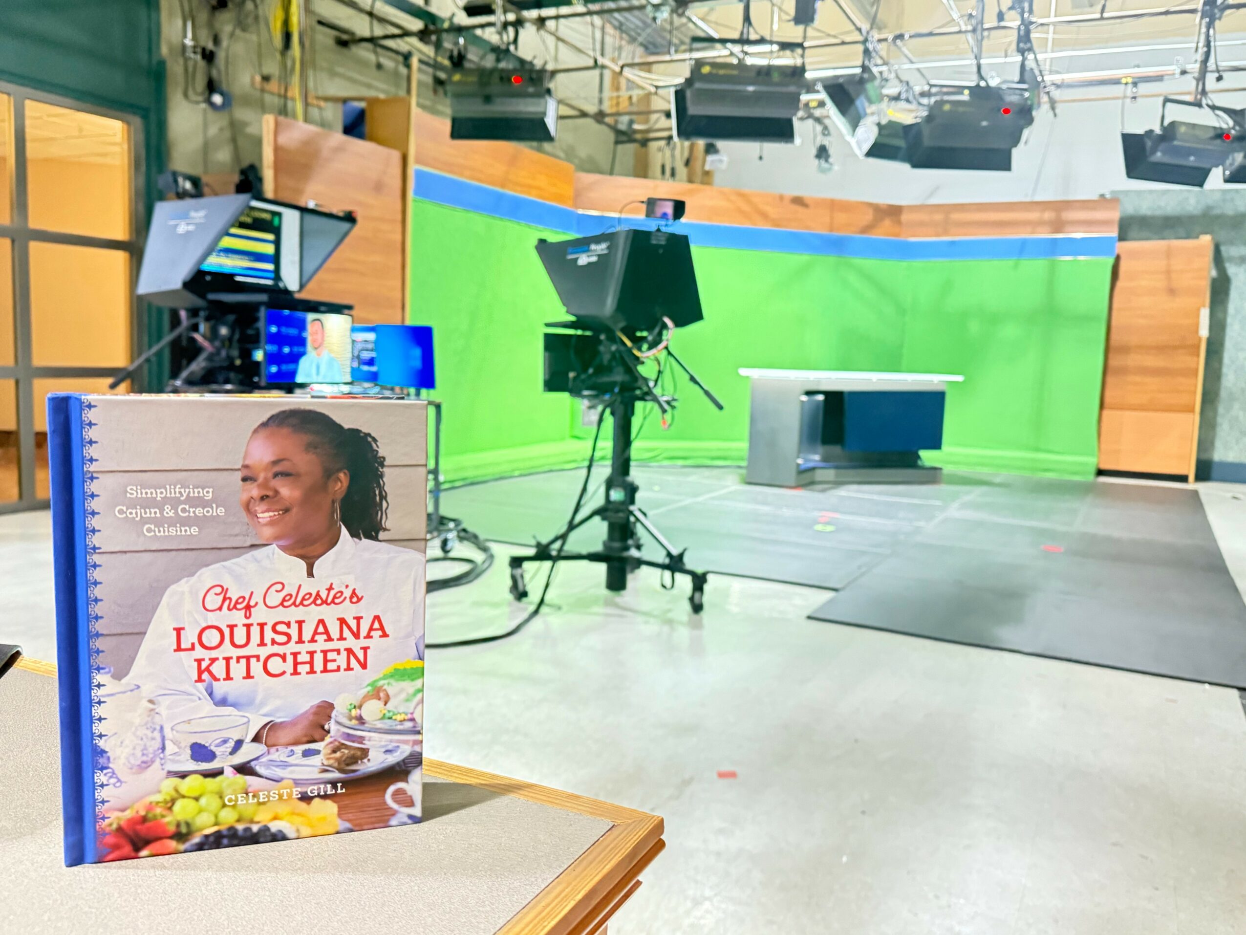 Television studio with green screen, camera, and desk, with a promotional sign for Louisiana Kitchen in foreground.