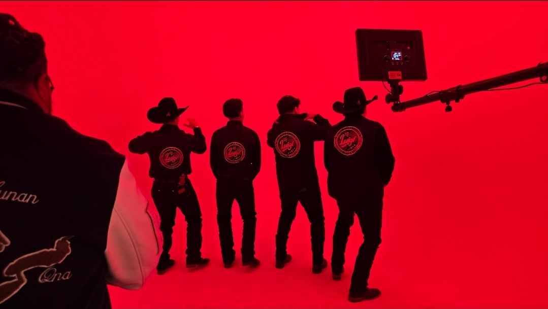 Five people stand in front of a red background, with one person in the foreground and a camera crane on the right.