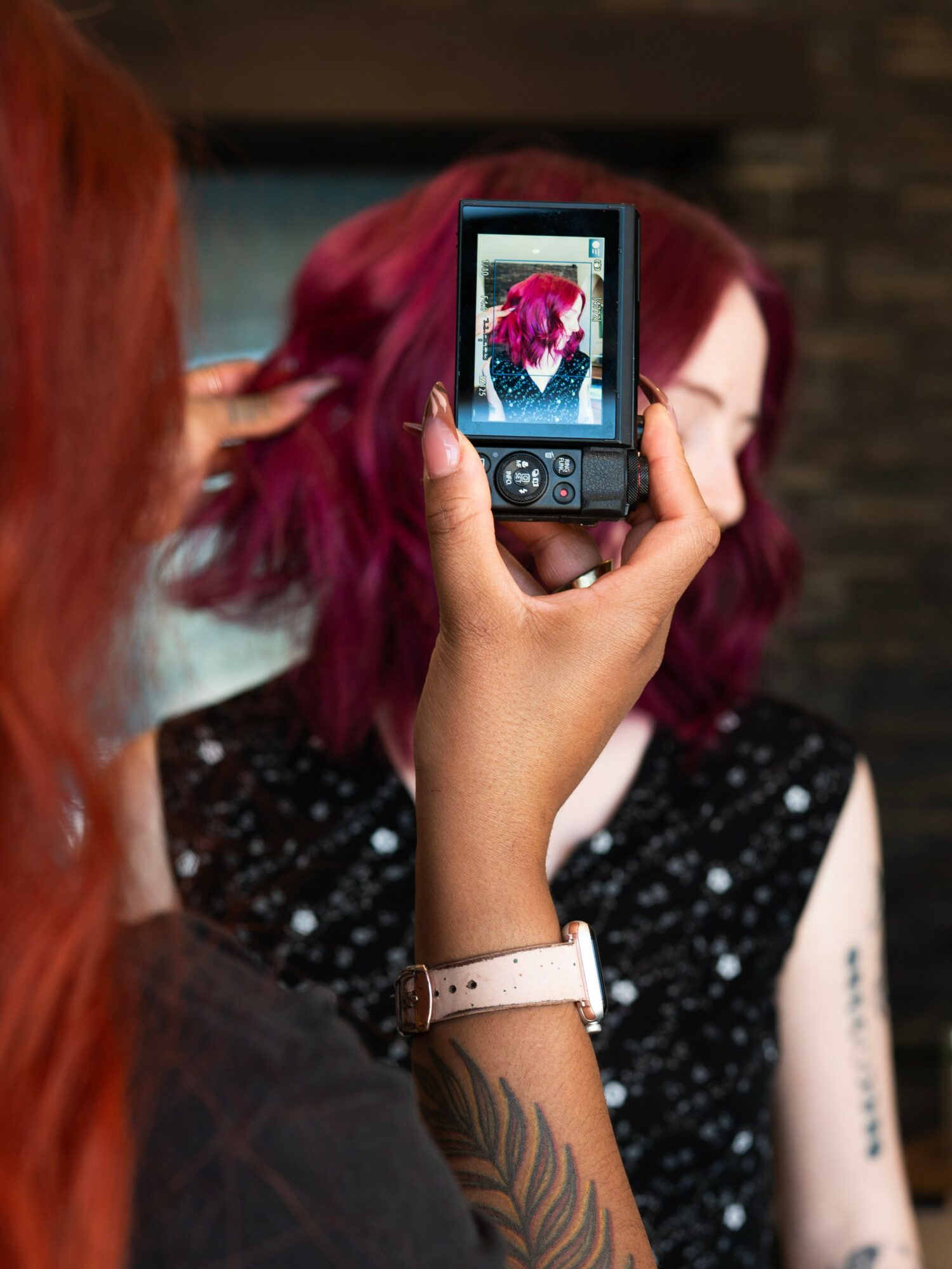 Person taking a photo of a woman with red and purple hair using a digital camera, indoors.