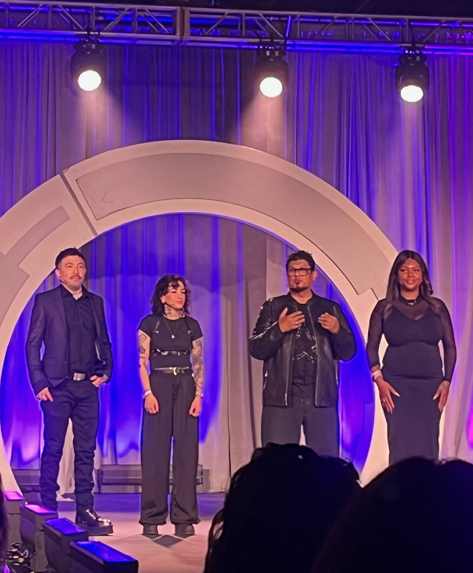 Four people stand on a stage with purple lighting and a large circular arch behind them.