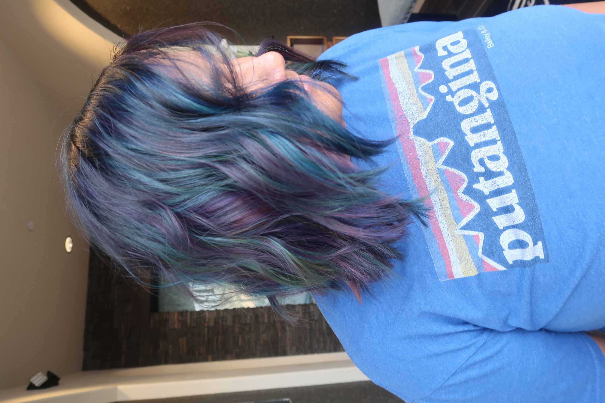 Person with multicolored hair, wearing a blue shirt with a logo, facing sideways in a room.