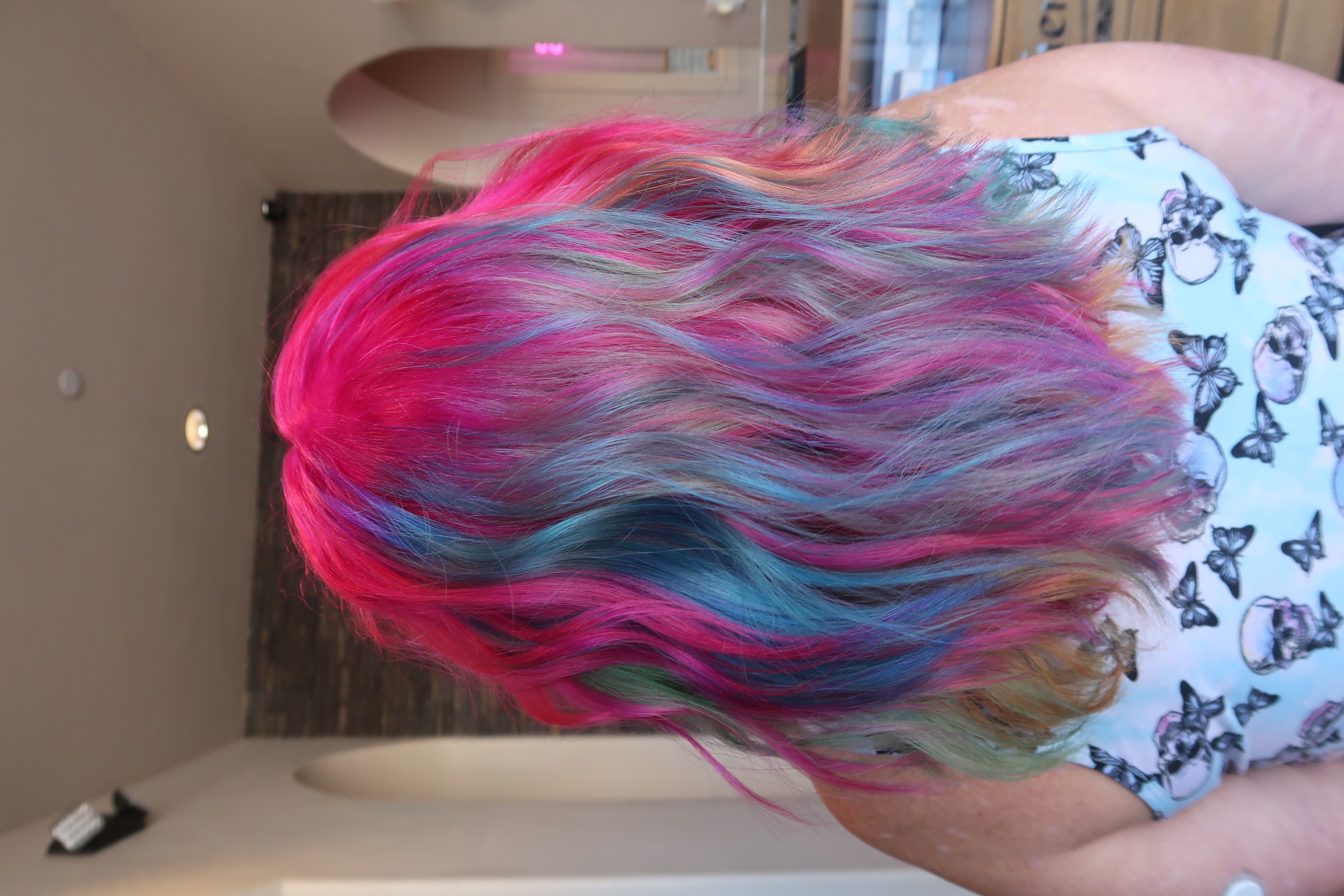 Back view of a person with multicolored, wavy hair in pink, blue, and blonde shades, wearing a butterfly-patterned top.