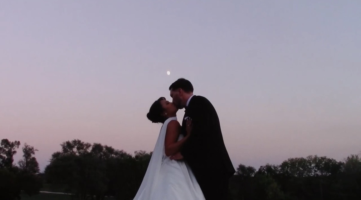 A couple in wedding attire kissing outdoors at dusk with trees and a visible moon in the sky.
