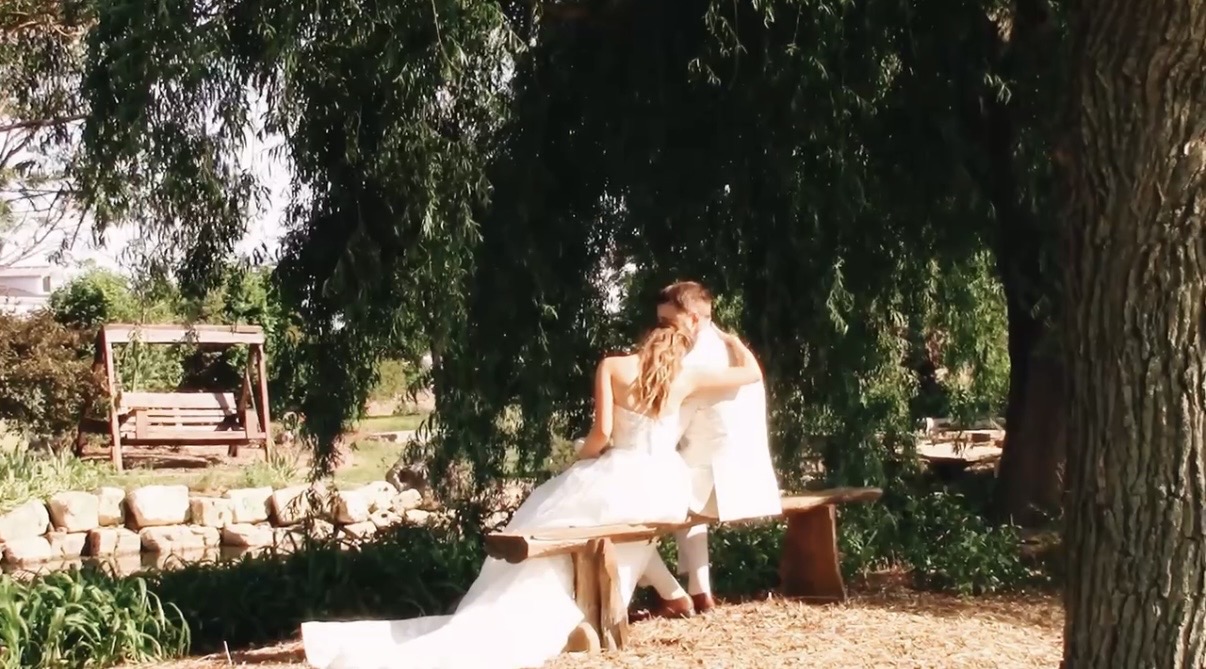 Two people in wedding attire embrace outdoors under trees, with a bench and stone wall in background.