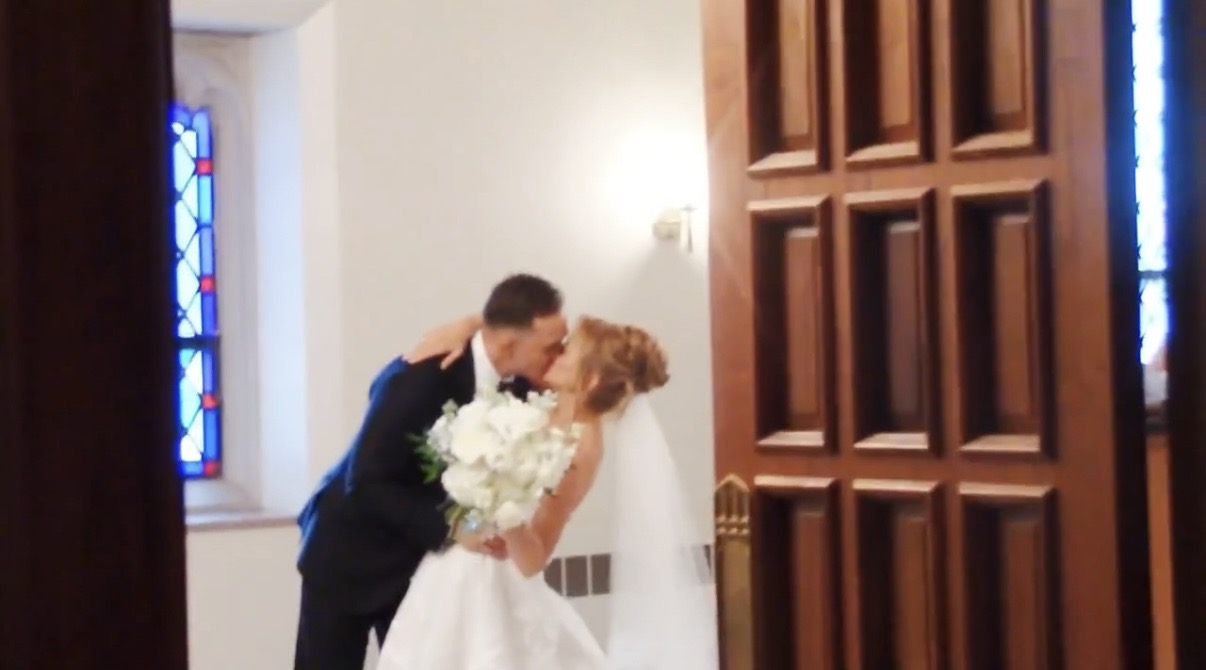 A bride and groom kiss inside a church, with stained glass window and wooden door visible.