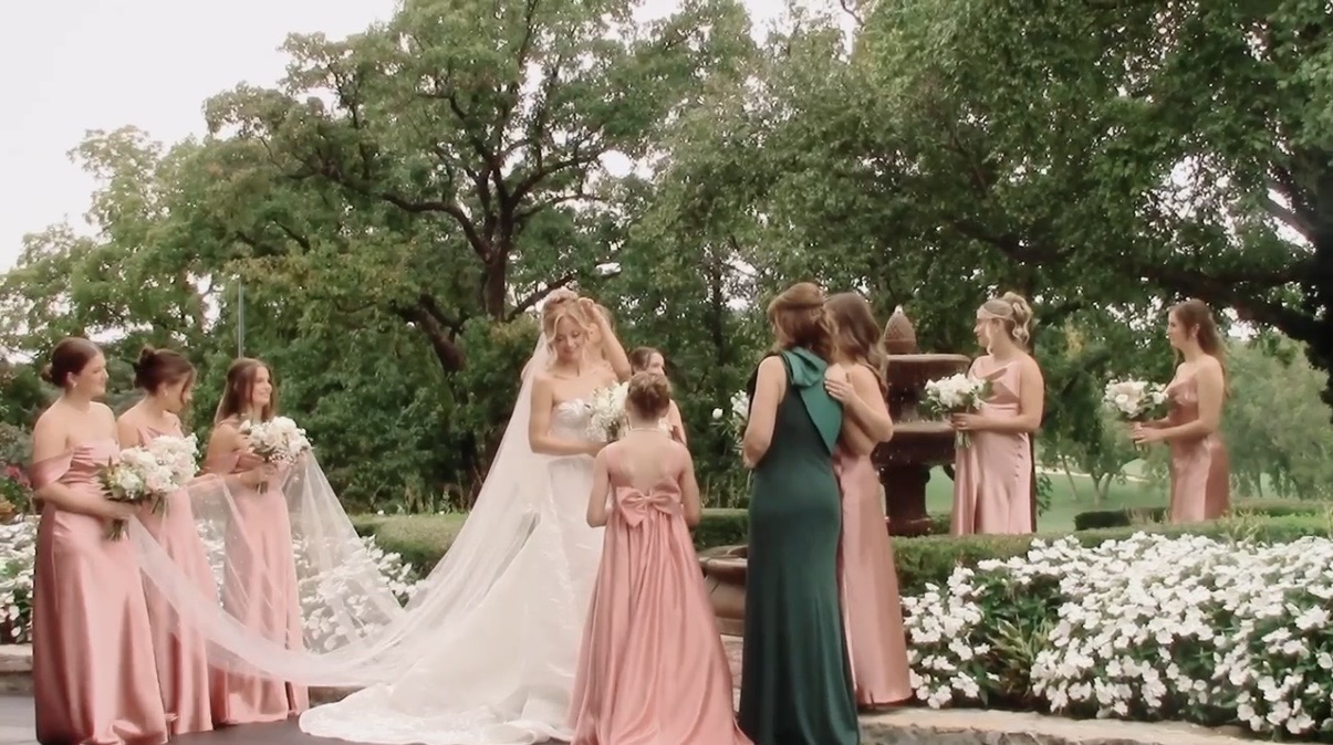 Wedding ceremony outdoors with bride, bridesmaids, and guests under trees, flowers, and greenery, during daytime.