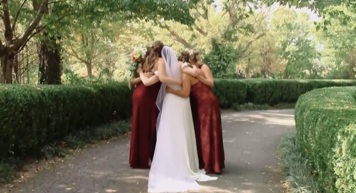 Three women in formal dresses hugging outdoors on a path with trees and bushes around.