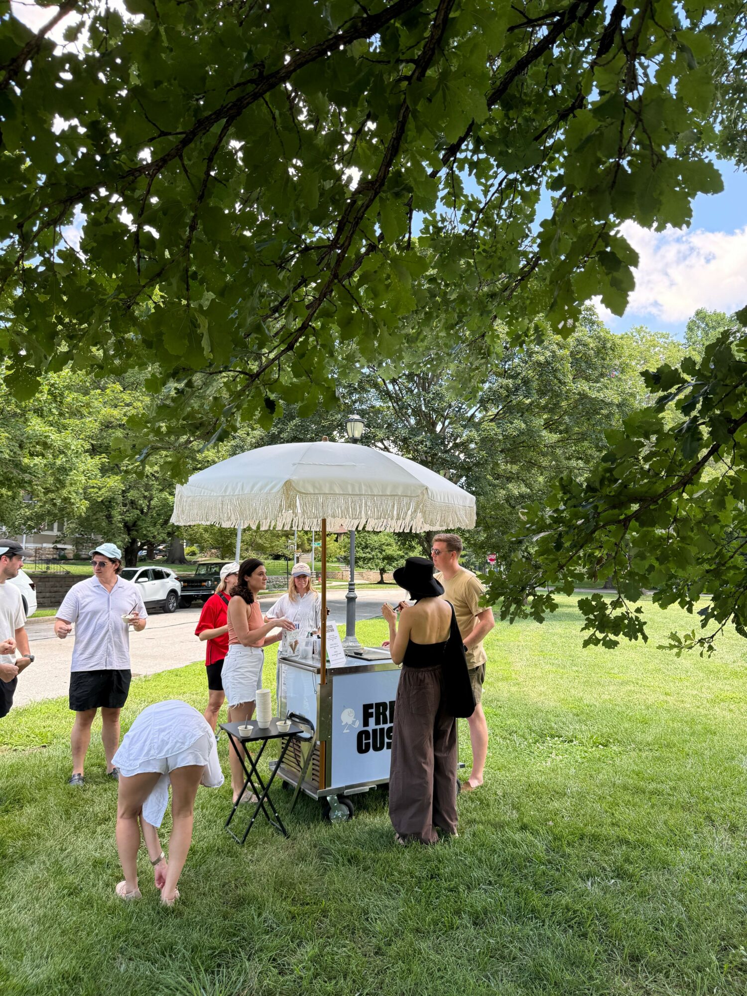 People walking outdoors near a sign, trees, and a grassy area, with a person holding an umbrella.
