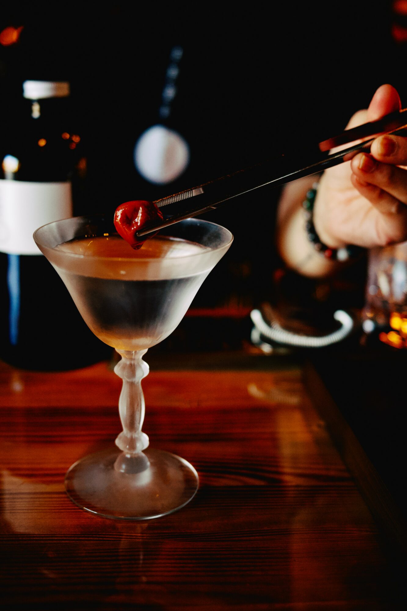 Person placing a cherry on a cocktail glass with a dark background, bar setting, and wooden surface.
