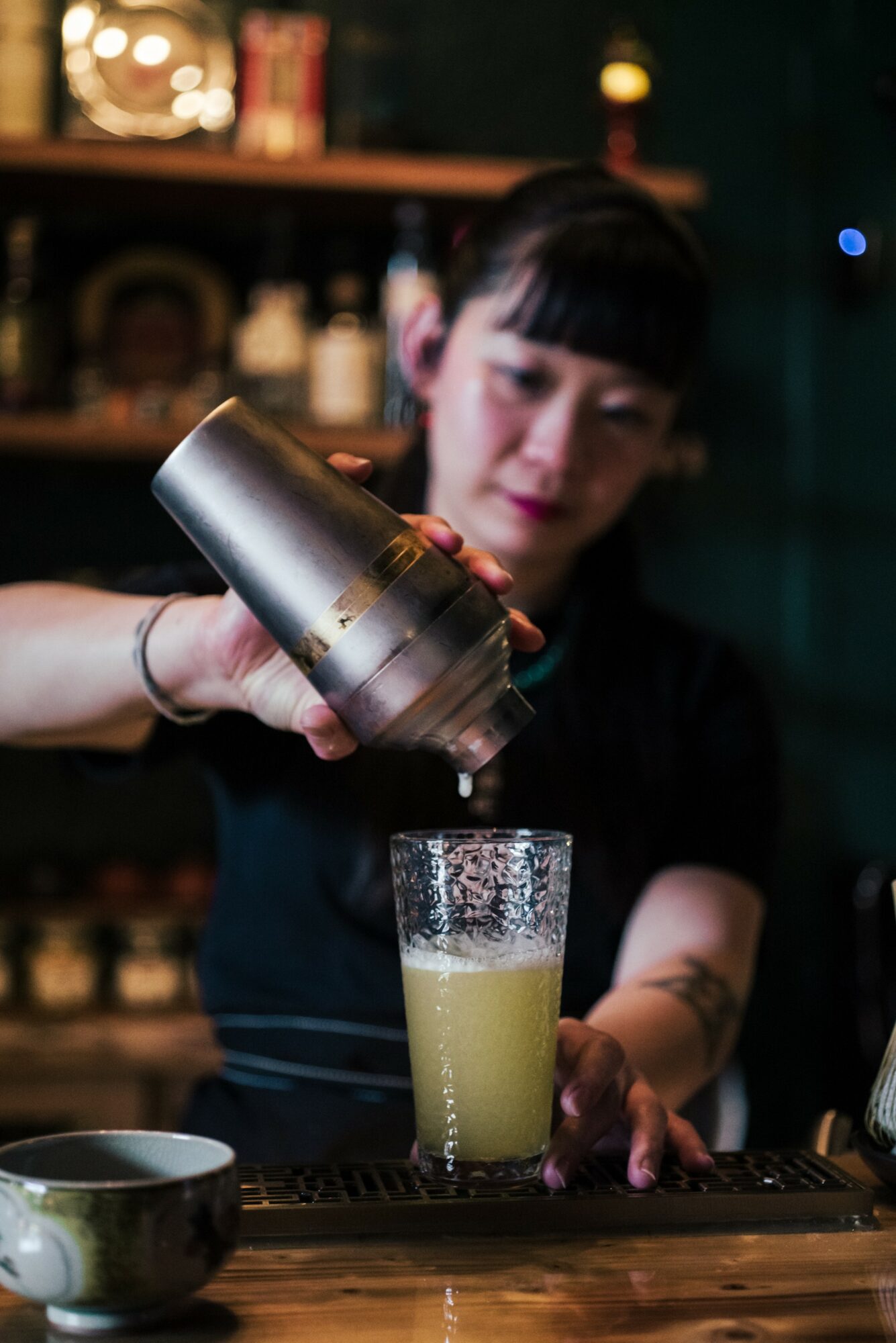 Person pouring a drink from a metal shaker into a glass, with a bar background.