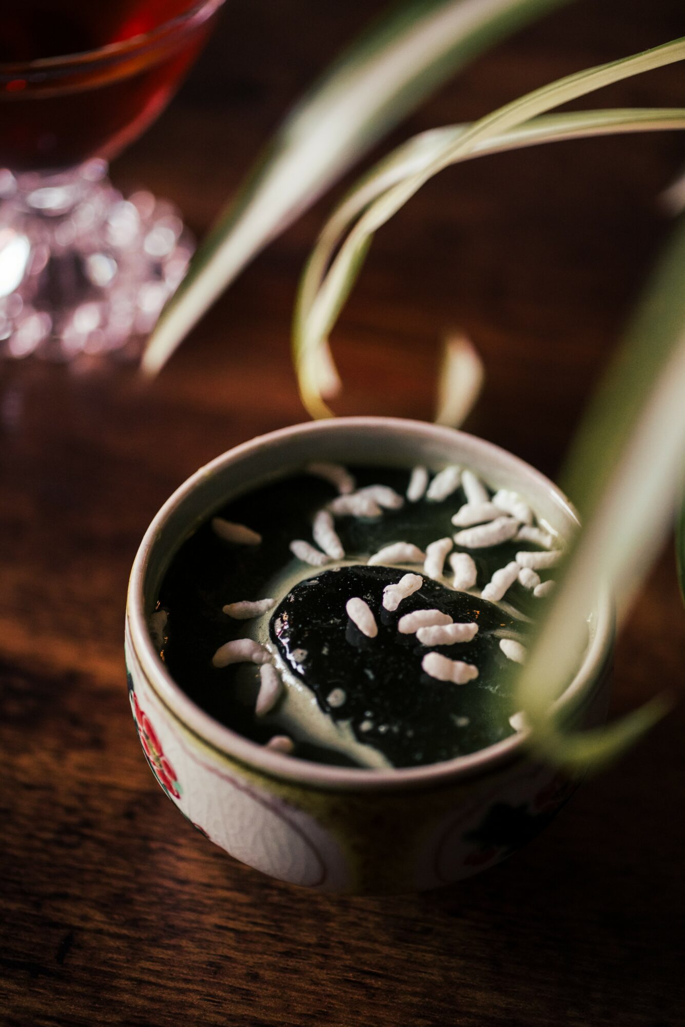 A cup of dark beverage topped with white seeds, with green leaves and a glass in the background, on a wooden surface.