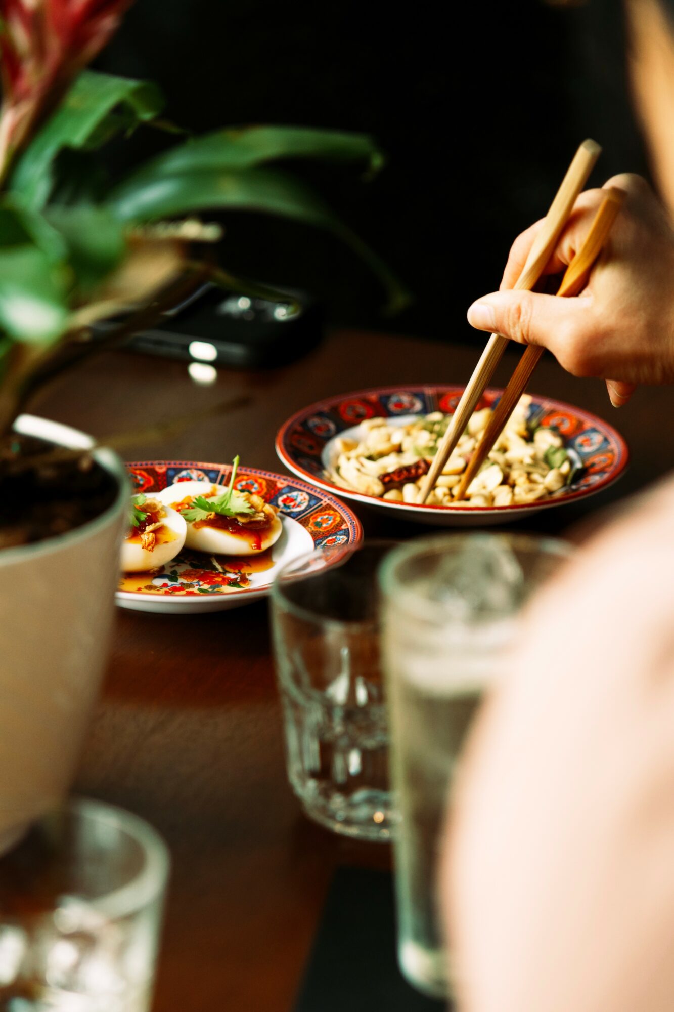 Person using chopsticks to eat from a bowl of food, with plates and glasses on a table.