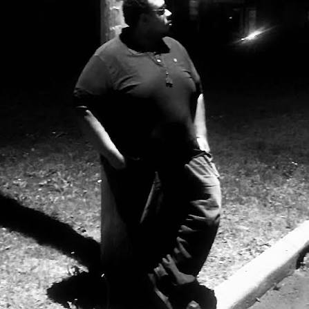 Person standing outdoors at night with hands in pockets, looking to the side, illuminated by a nearby light source.