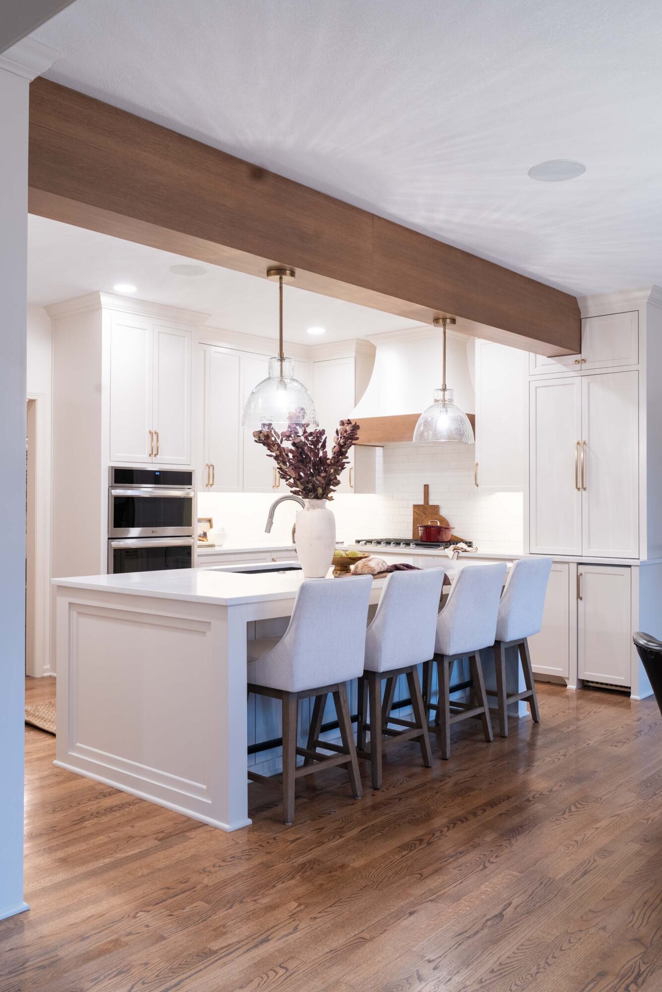 Kitchen with white cabinets, island with four chairs, pendant lights, and a vase with purple flowers, hardwood floor.