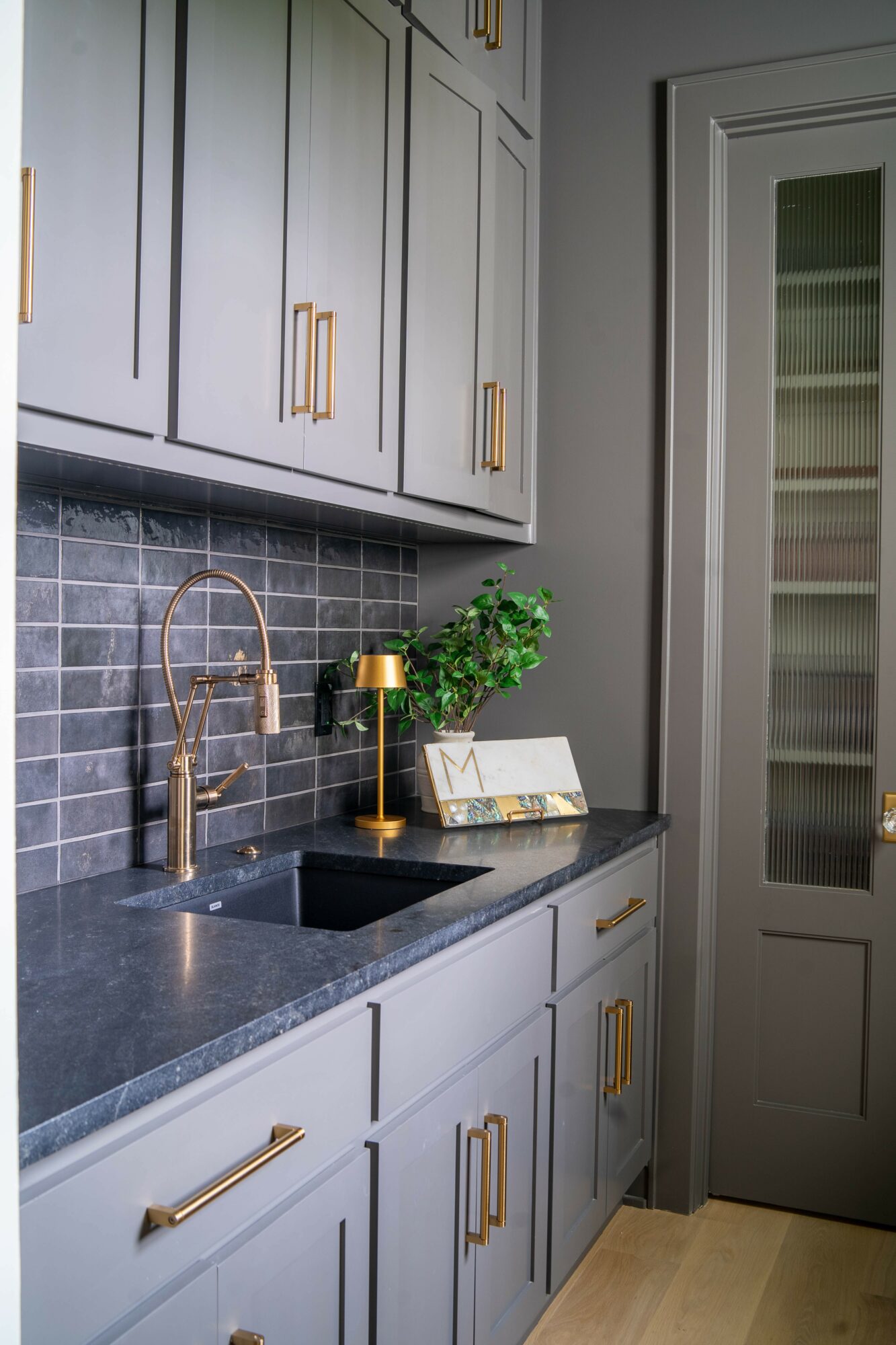 Kitchen with gray cabinets, black countertop, gold hardware, and a black backsplash with gray tiles. A gold faucet and a small plant are on the counter.