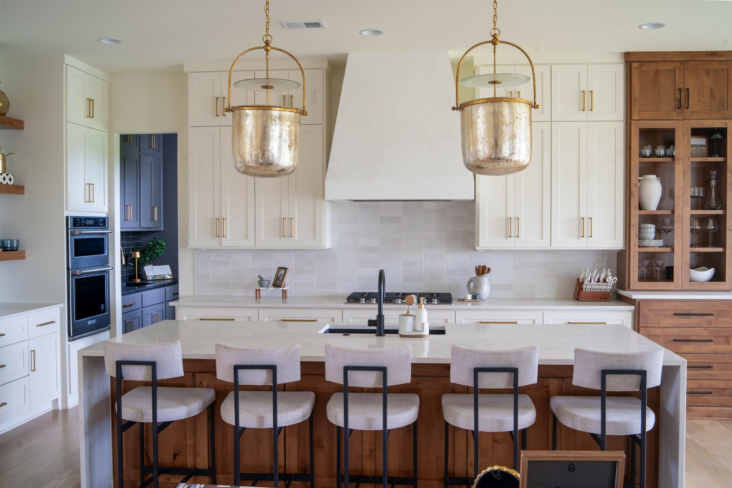 Modern kitchen with white cabinets, a large island with six chairs, and two hanging pendant lights, with a wooden cabinet on the right.