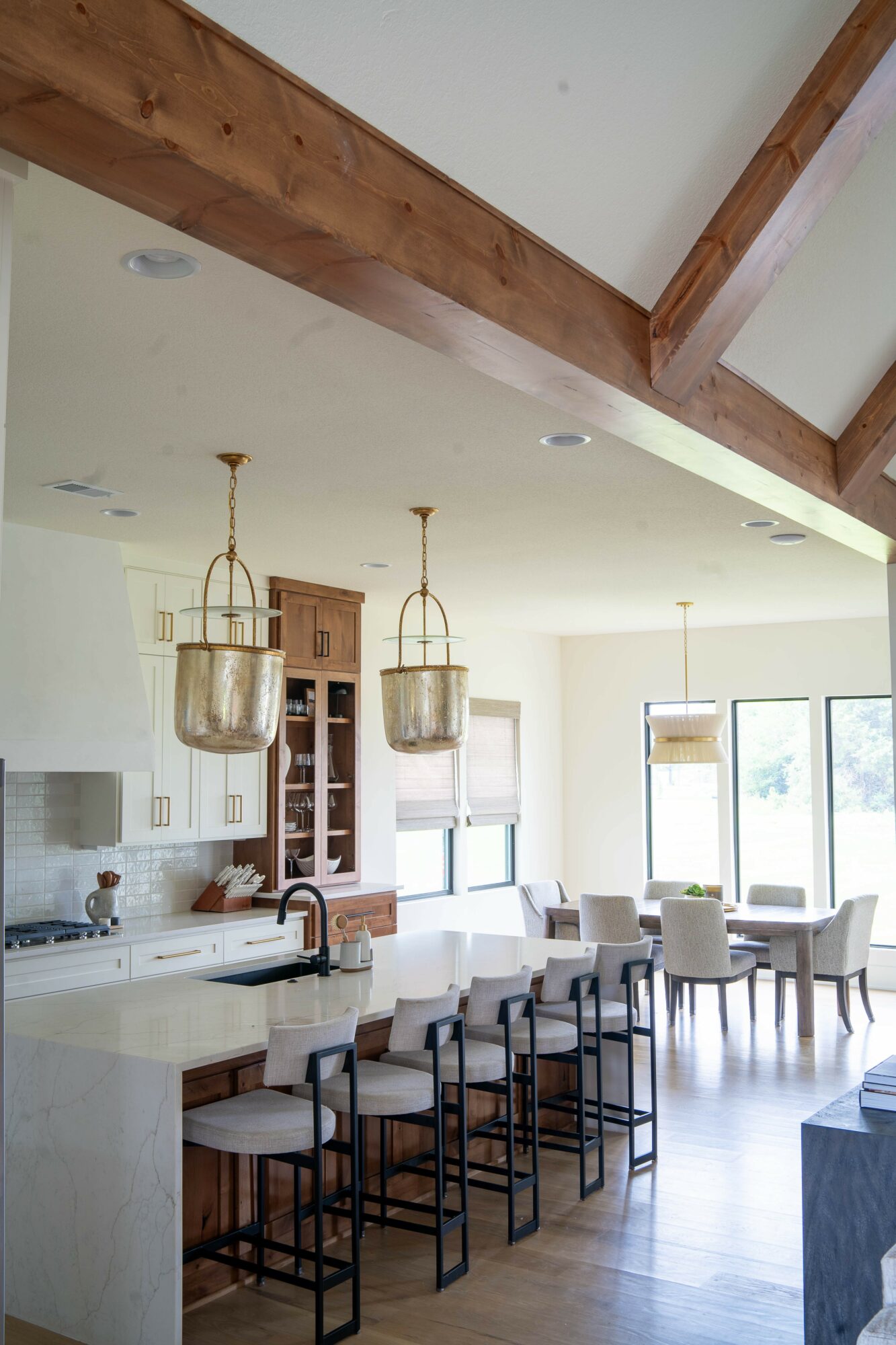 Open kitchen and dining area with white walls, wooden beams, and large windows, featuring a kitchen island with bar stools.