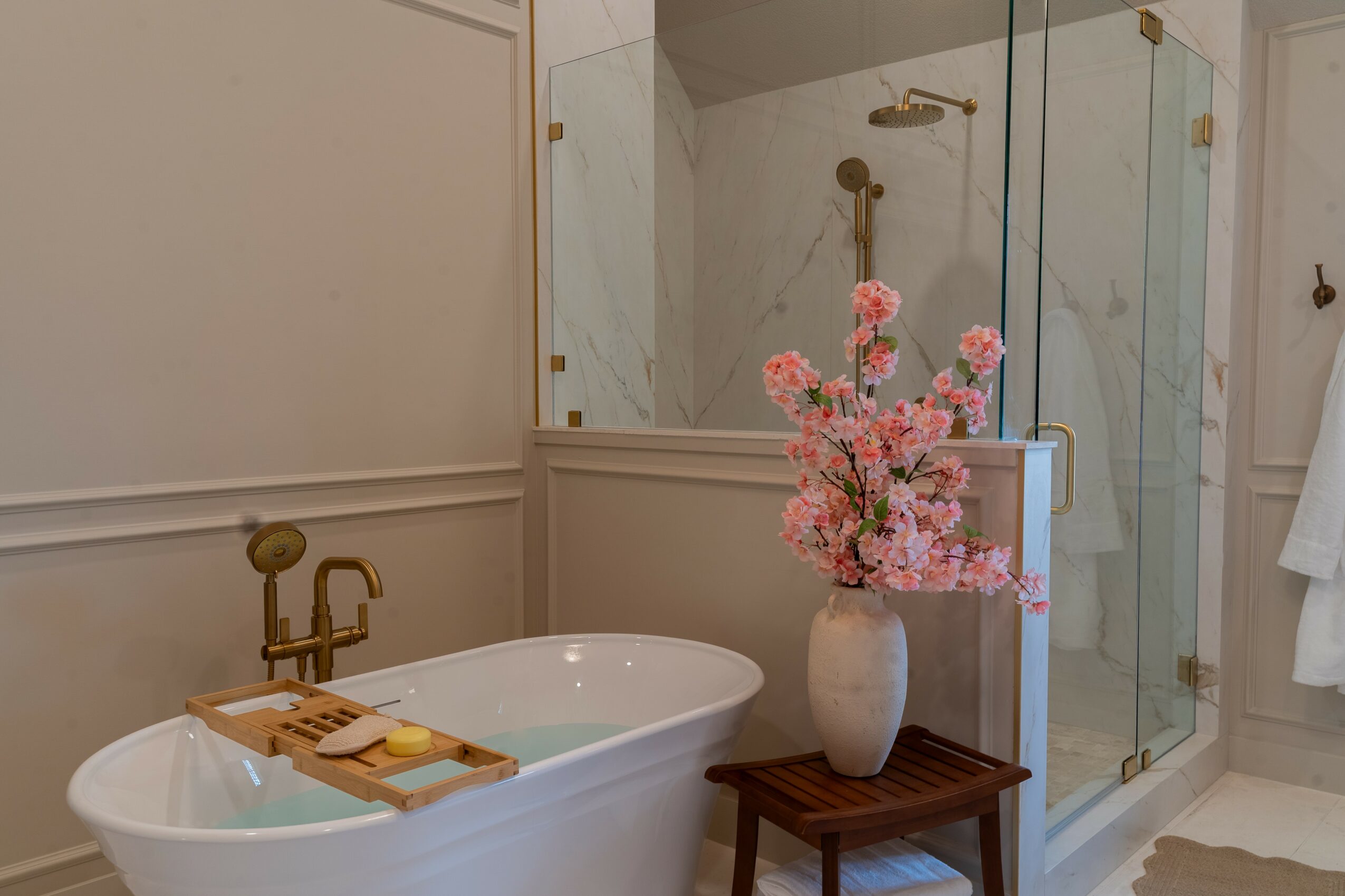 Bathroom with bathtub, glass shower, pink flowers in a vase, and a wooden stool.