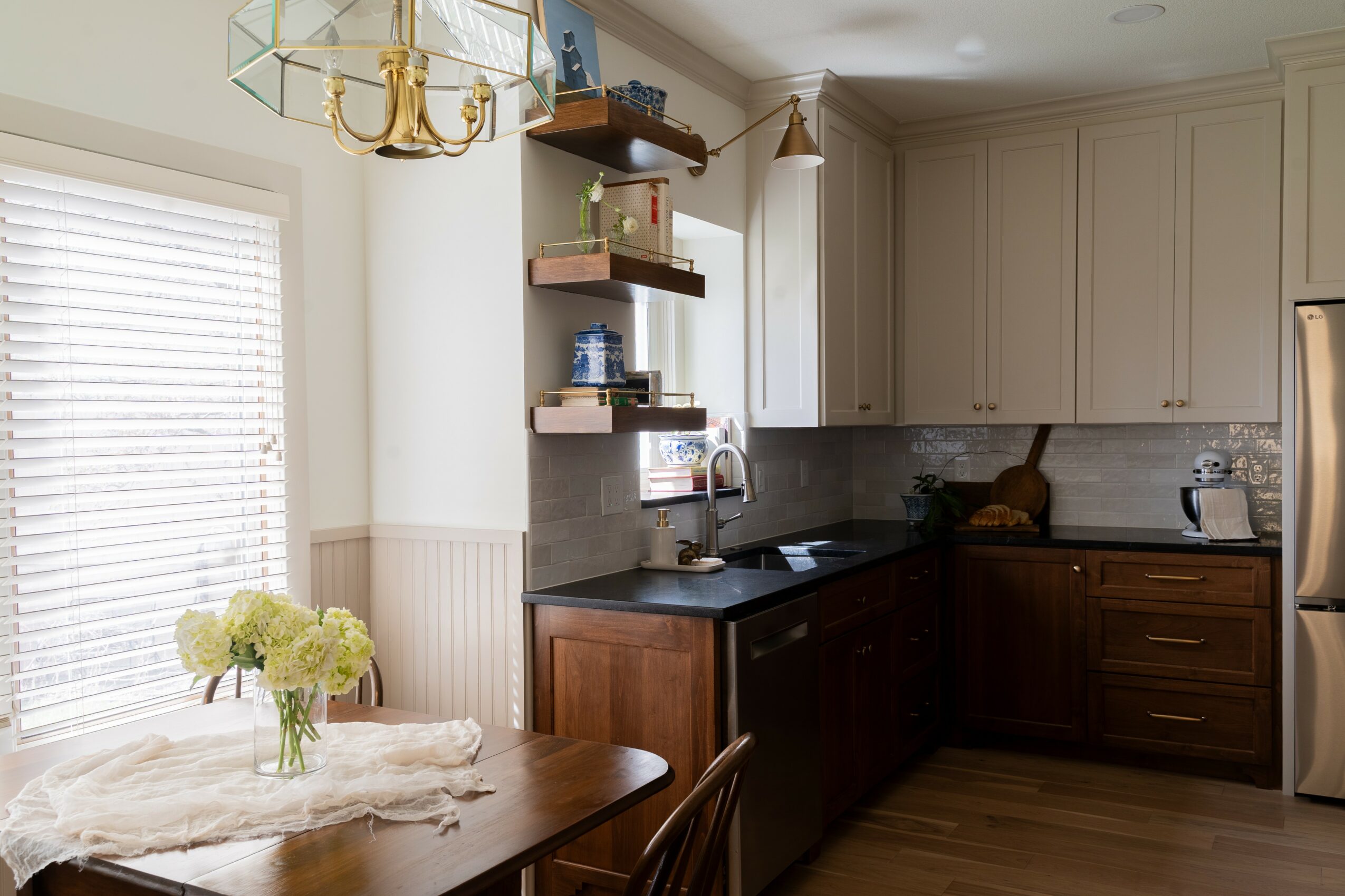 Kitchen with beige cabinets, dark countertop, and a window with blinds; dining table with flowers in a vase.
