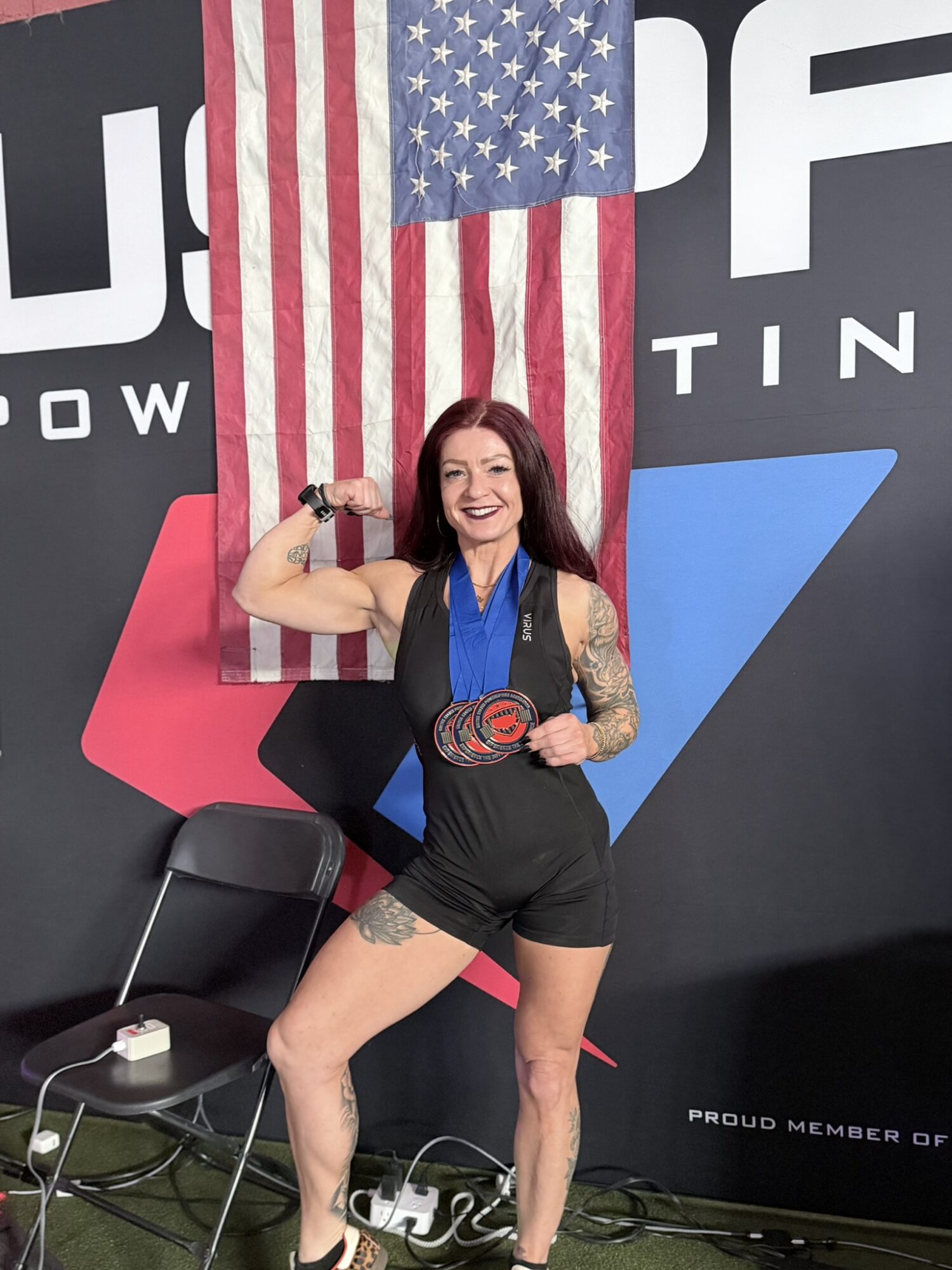 Woman with tattoos flexing arm, smiling, standing in front of American flag and a black wall with text, wearing black shorts and a medal.