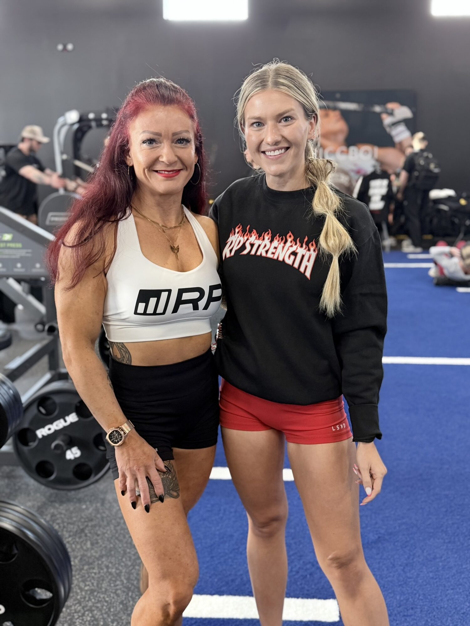 Two women smiling at a gym, one with red hair and the other with blonde hair, standing close together.