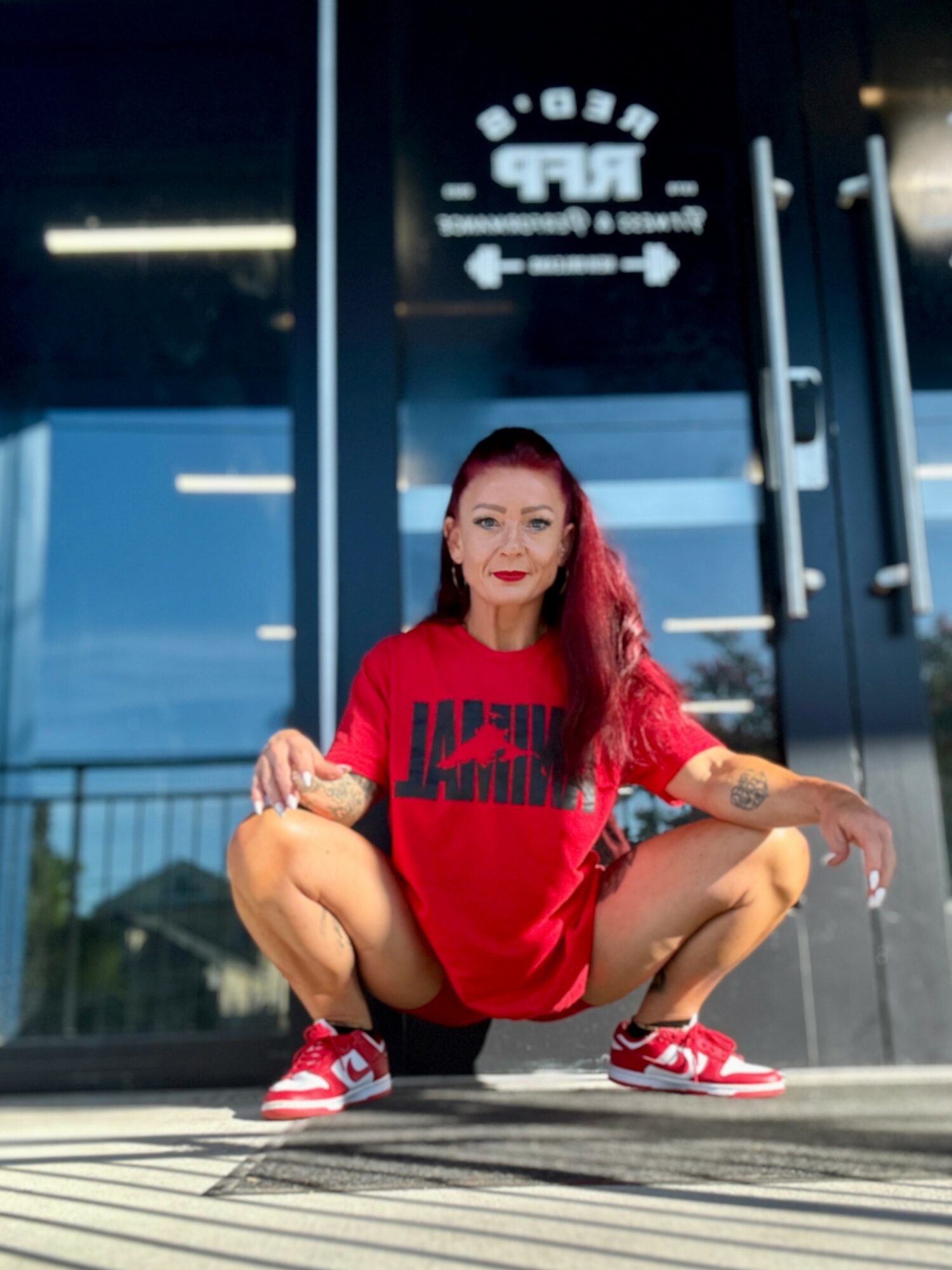 Woman with red hair squatting outside a building, wearing a red shirt and sneakers, smiling at the camera.