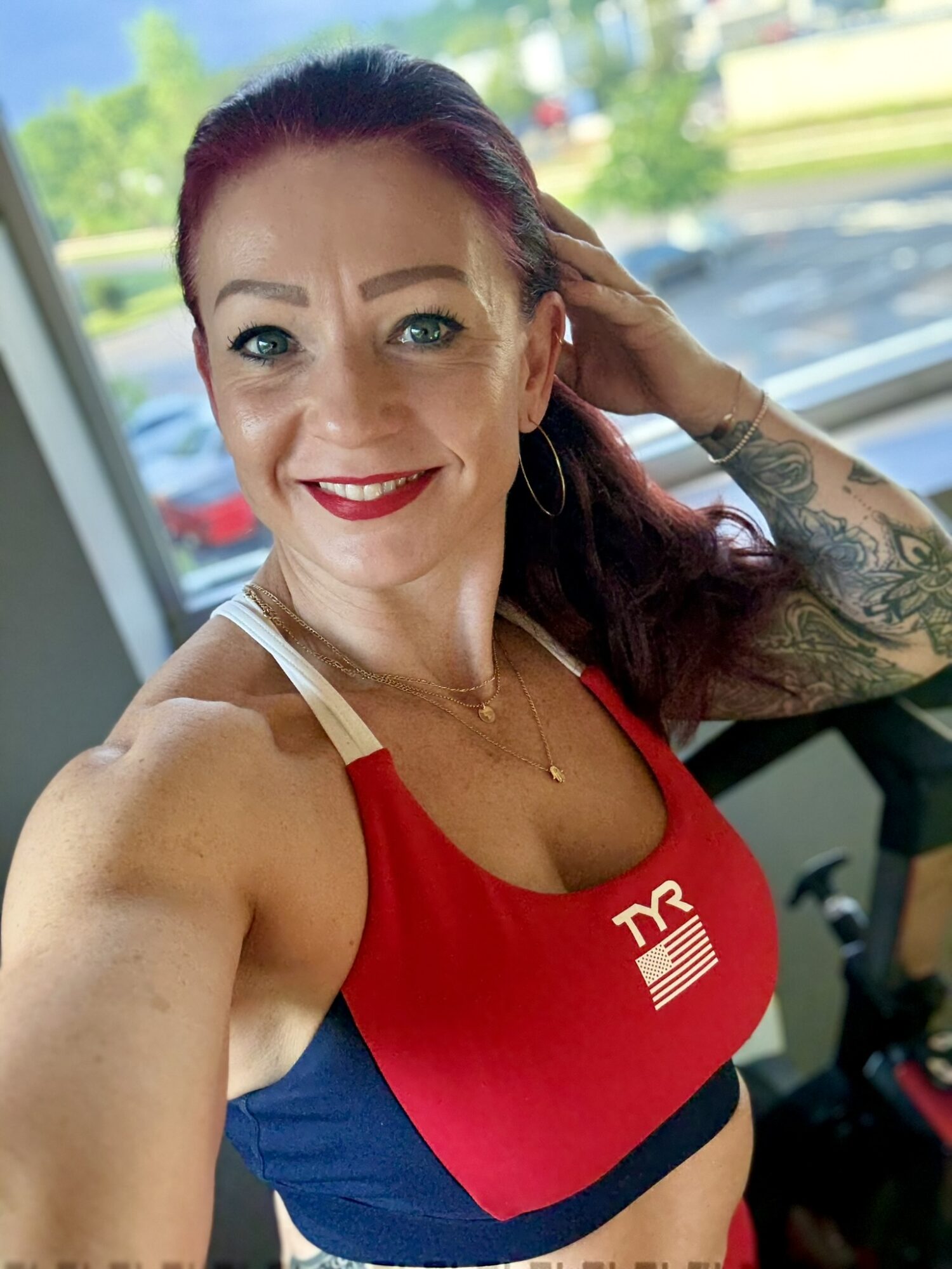 Woman with red hair and tattoos smiling, wearing a red and blue sports top, indoors with windows and trees outside.