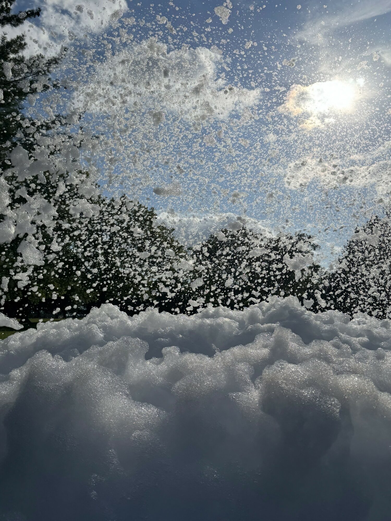 Snow-covered ground with snow falling and a bright sun in a partly cloudy sky.