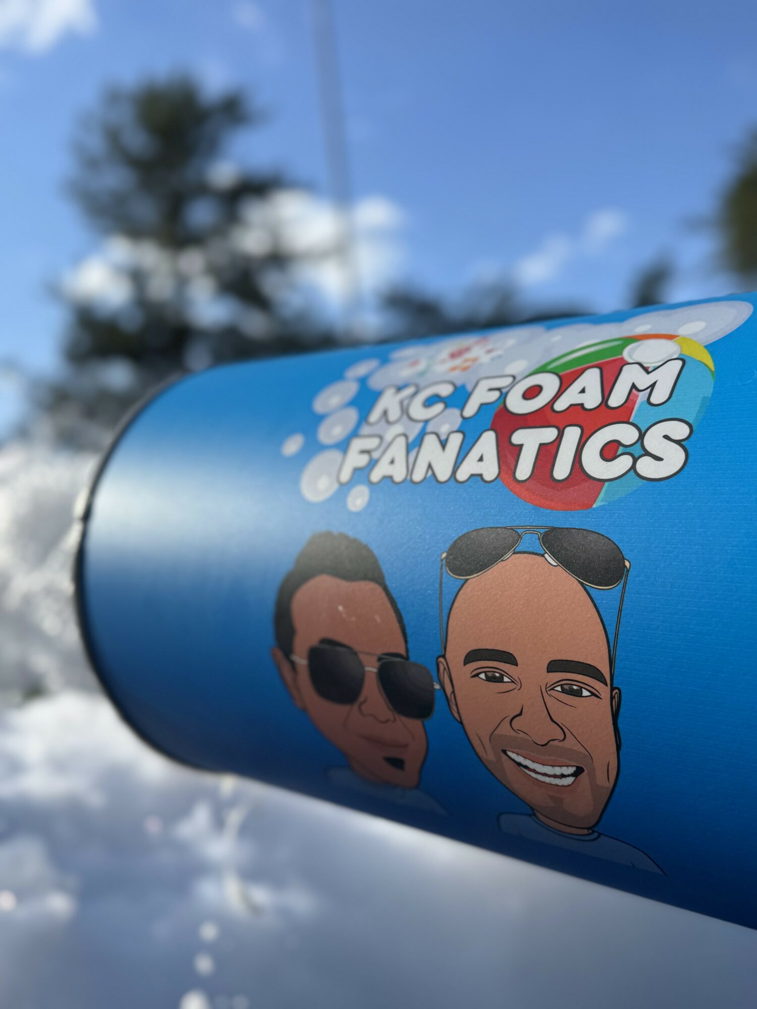 Blue cylindrical object with a logo and cartoon faces, outdoors with trees and sky in background.