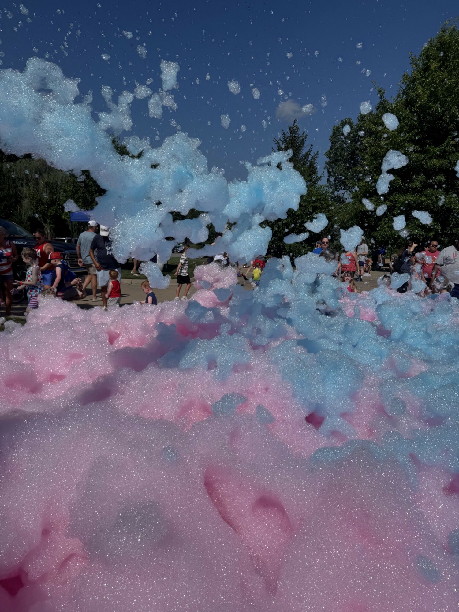 People outdoors with foam and bubbles, trees in background, blue sky, foam on ground, some pink foam visible.