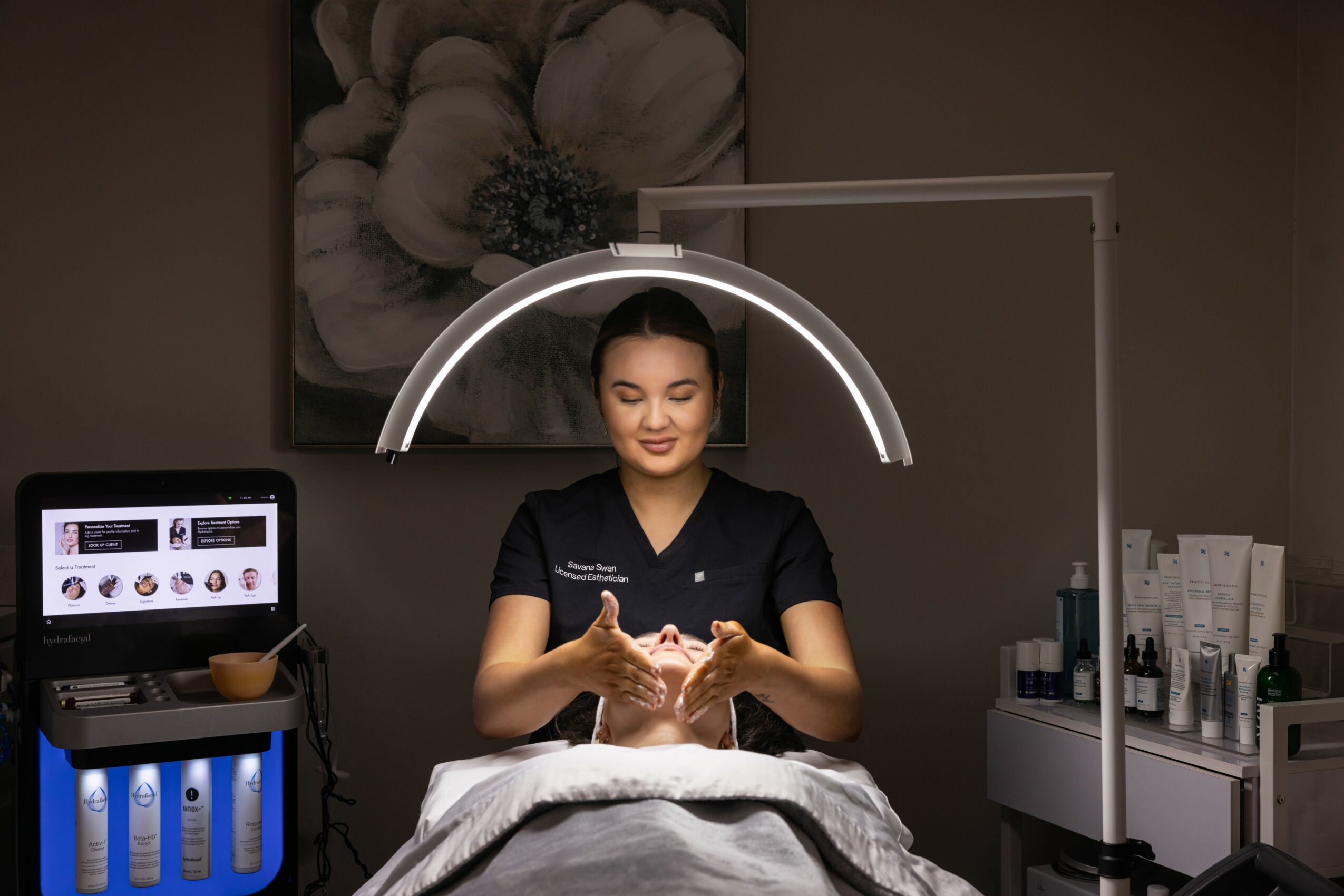 A woman receives a facial treatment under a circular light, with skincare products nearby, in a spa or clinic setting.