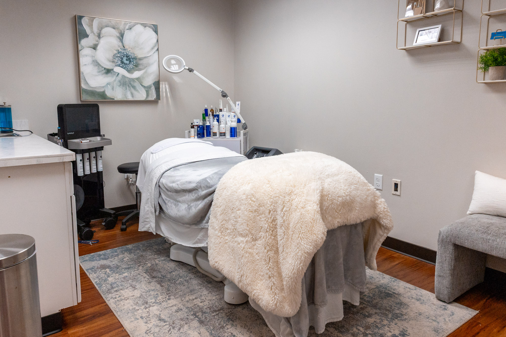 A medical or spa treatment room with a bed covered in a white blanket, medical equipment, and a gray armchair.