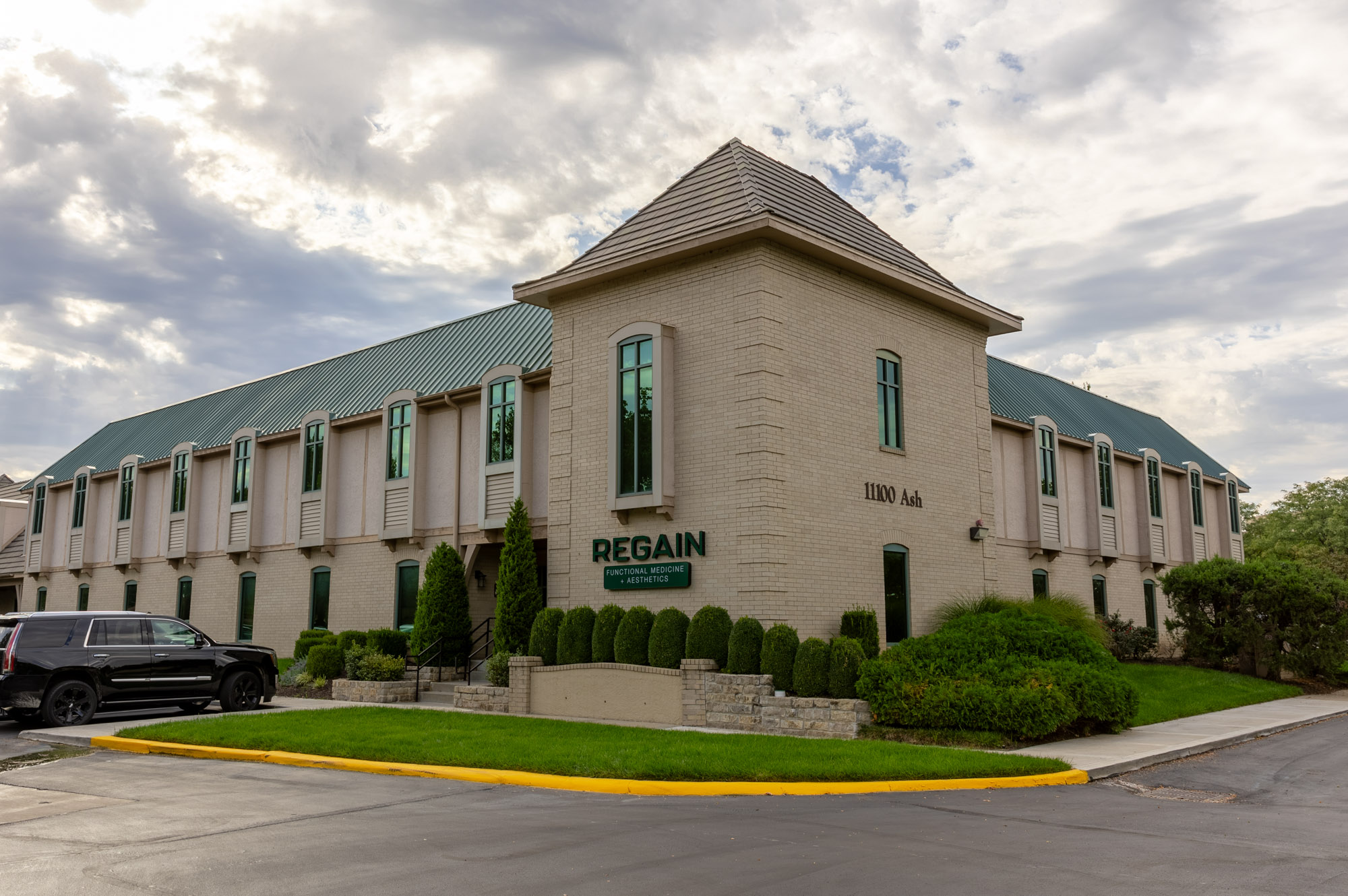 Large building with a rounded corner, multiple windows, and a sign that reads 'REGAIN'. Green bushes and trees surround the building.