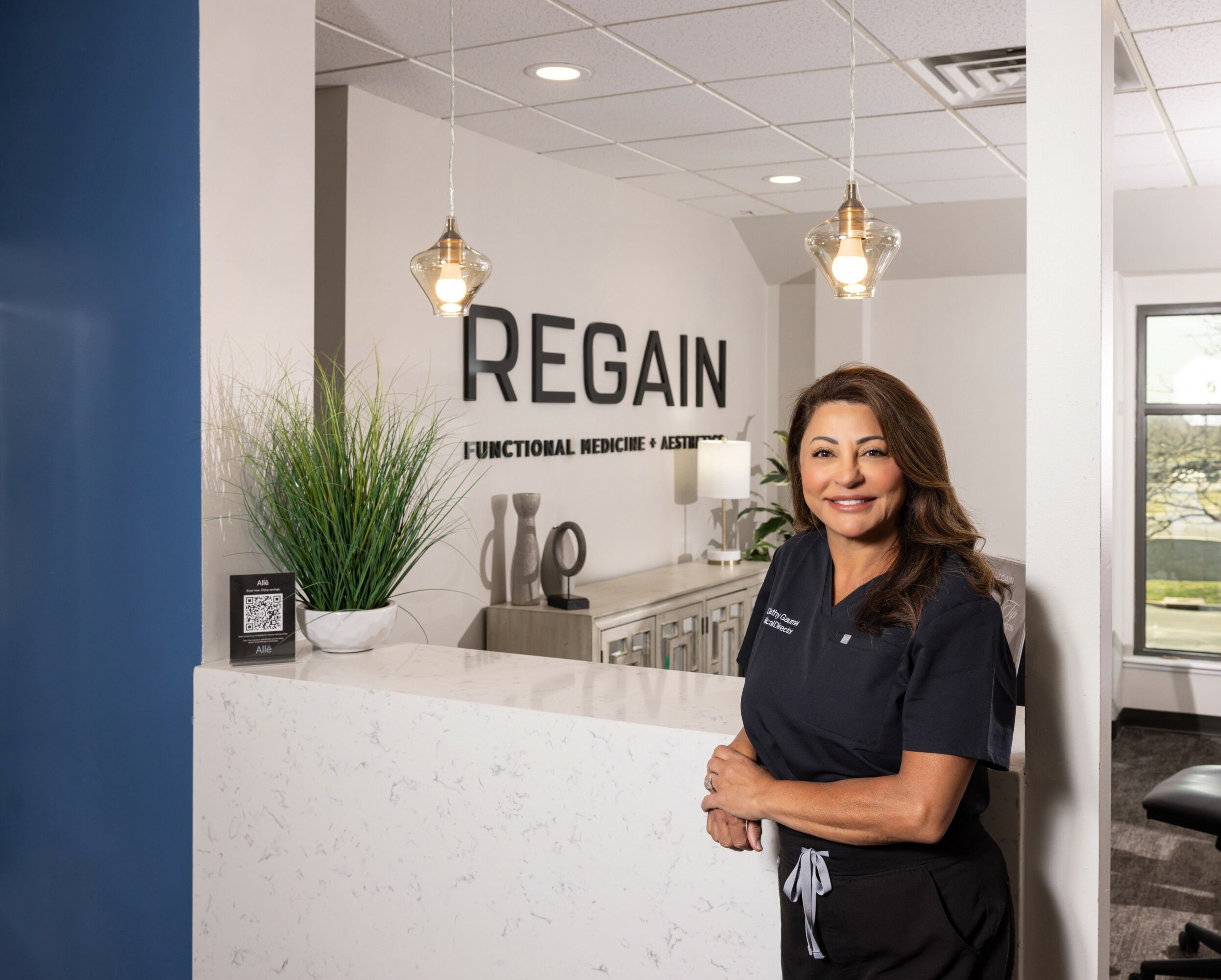 Woman in medical scrubs standing at reception desk in clinic with 'REGAIN' sign behind her.