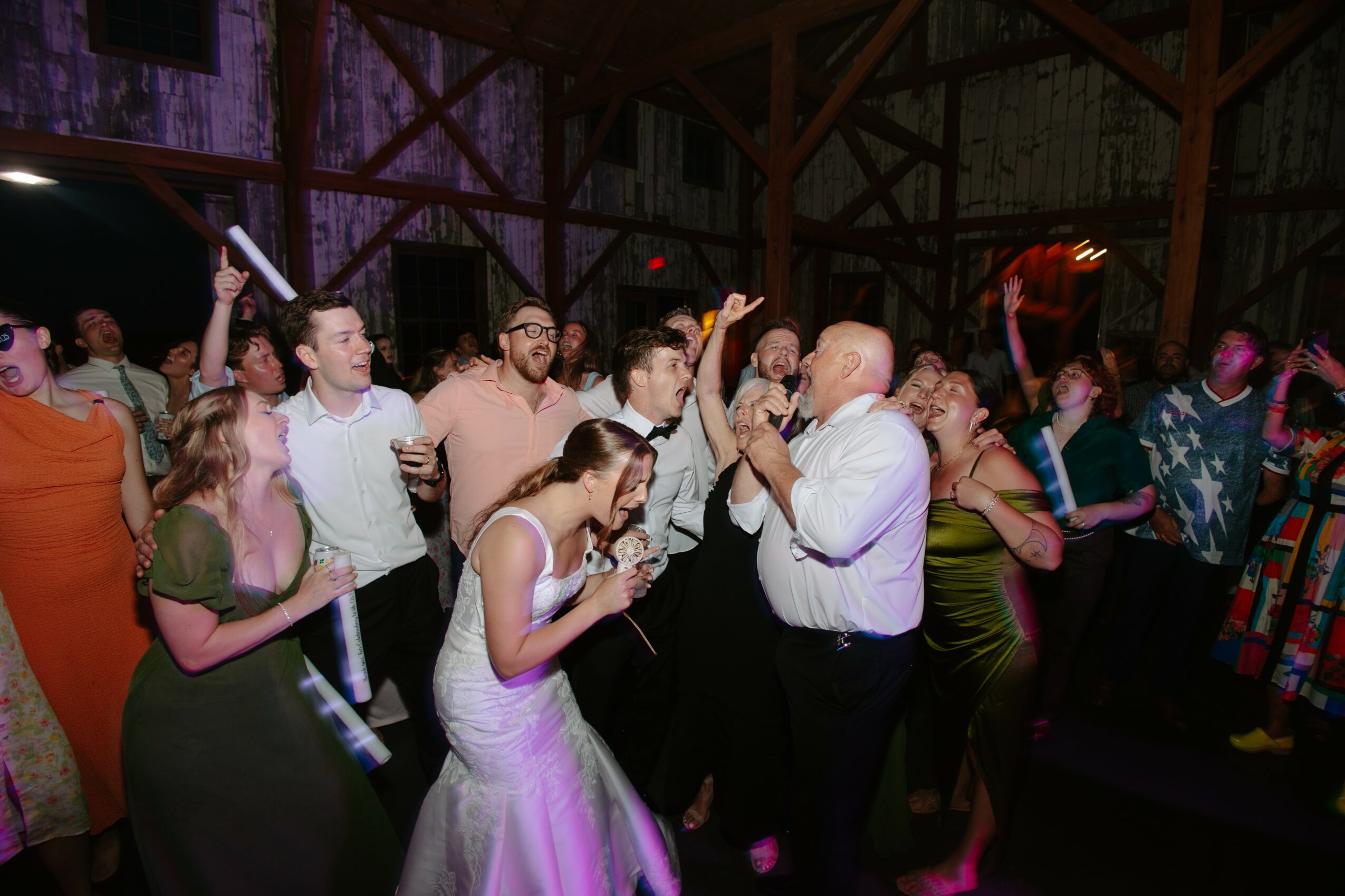 People dancing and celebrating in a rustic venue with wooden beams, some holding glow sticks, in a lively party scene.