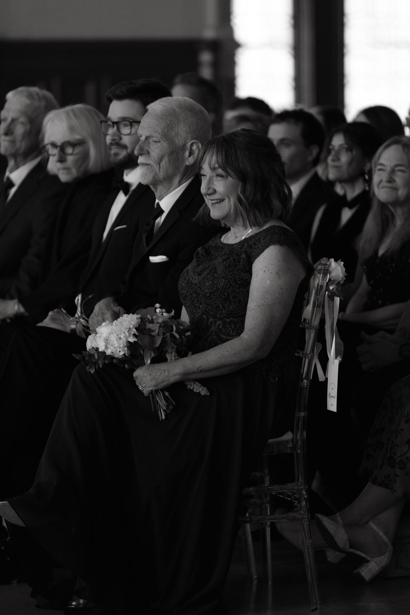 Group of people seated at an event, some smiling, dressed formally, holding flowers.
