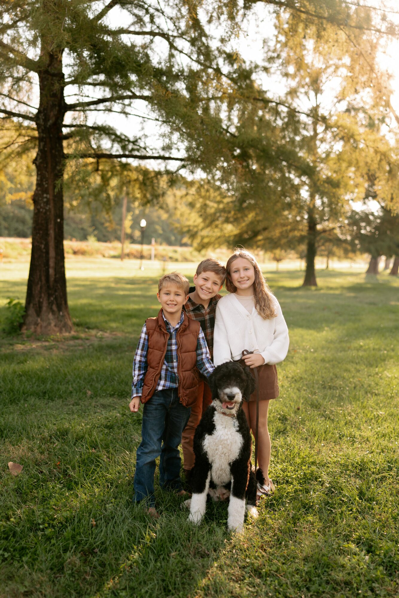 Three children and a dog standing outdoors in a park with trees and grass, smiling at the camera.
