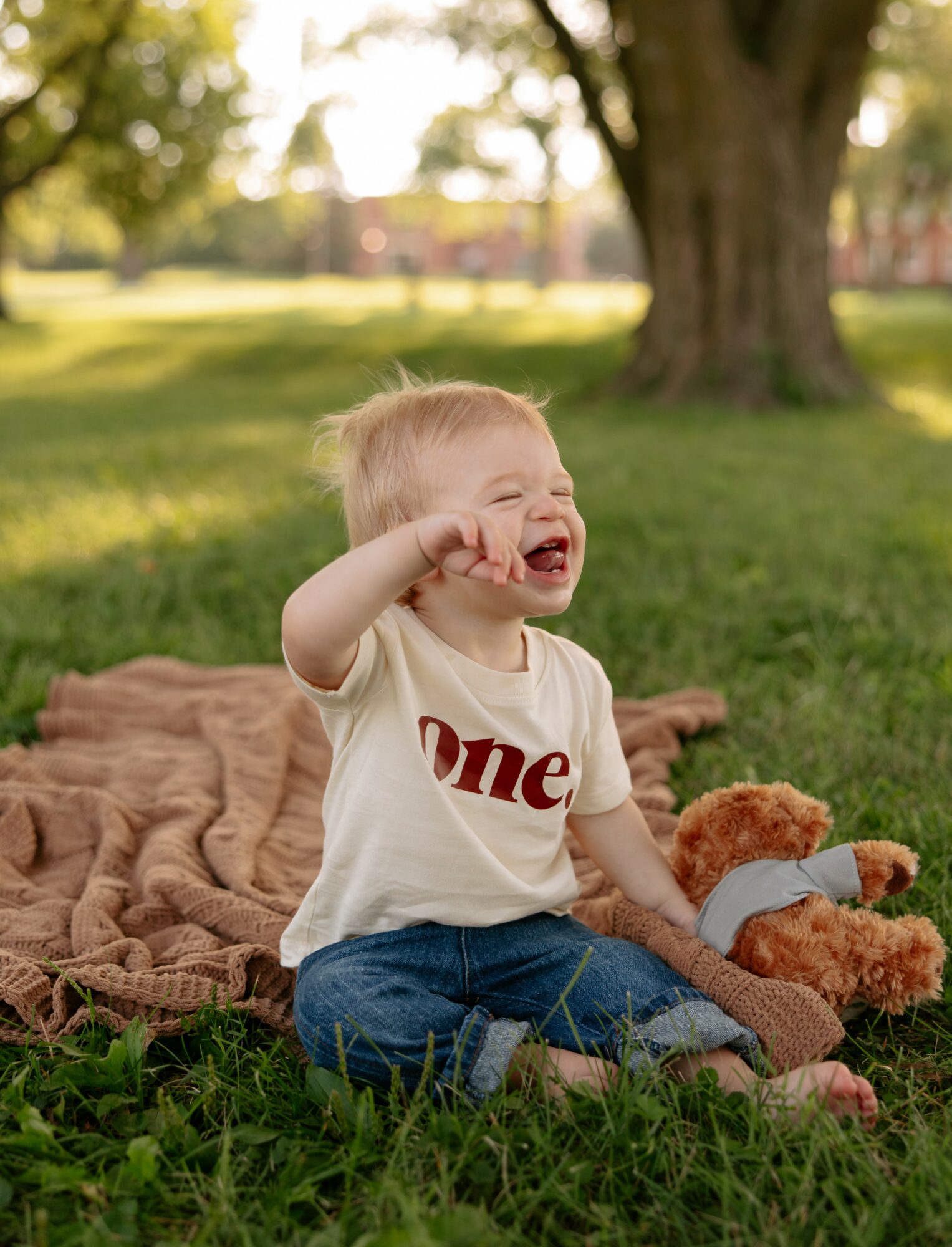 Child sitting on a blanket outdoors, laughing, with a teddy bear, trees, and sunlight in the background.