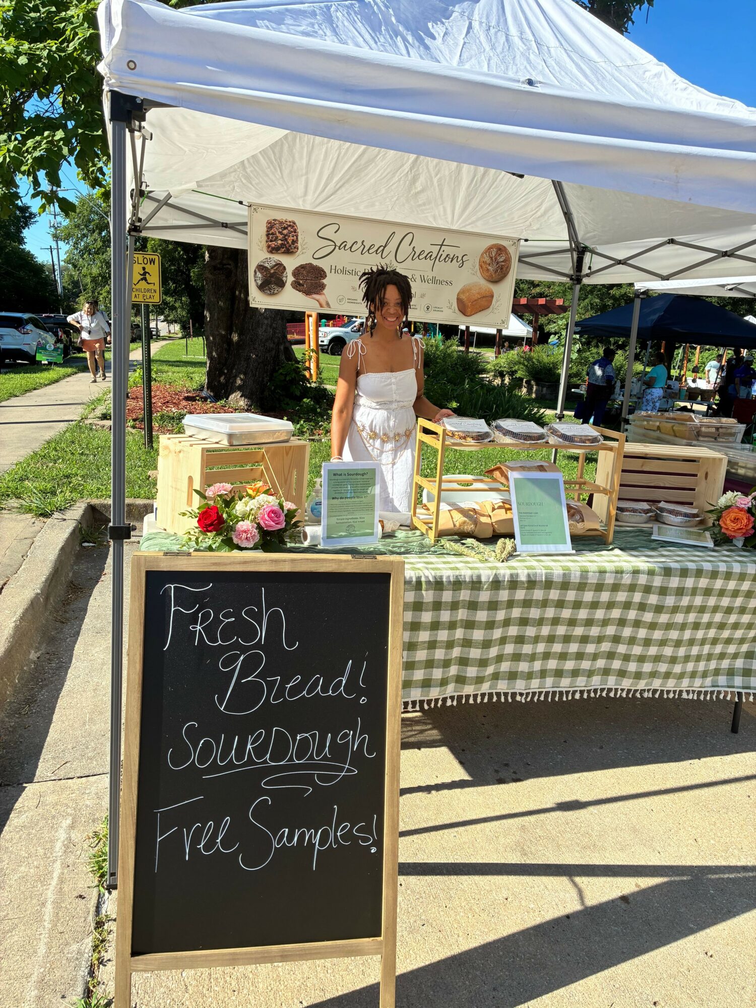 Woman at a booth with a chalkboard sign reading 'Fresh Beautiful Soups, Free Samples' at an outdoor market or fair.