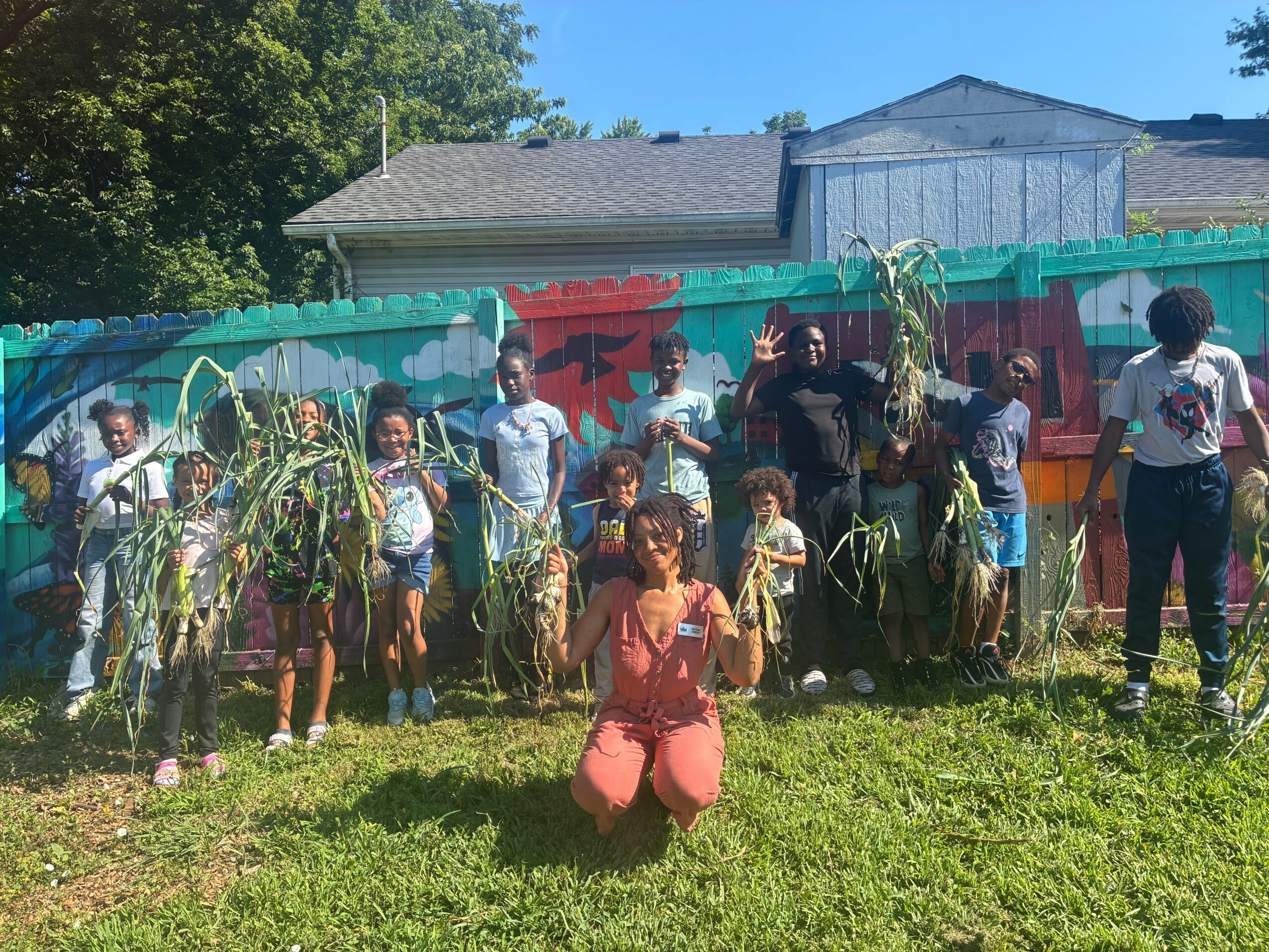 Group of children and an adult outdoors in front of a colorful mural, holding plants or branches, with a woman kneeling in front.