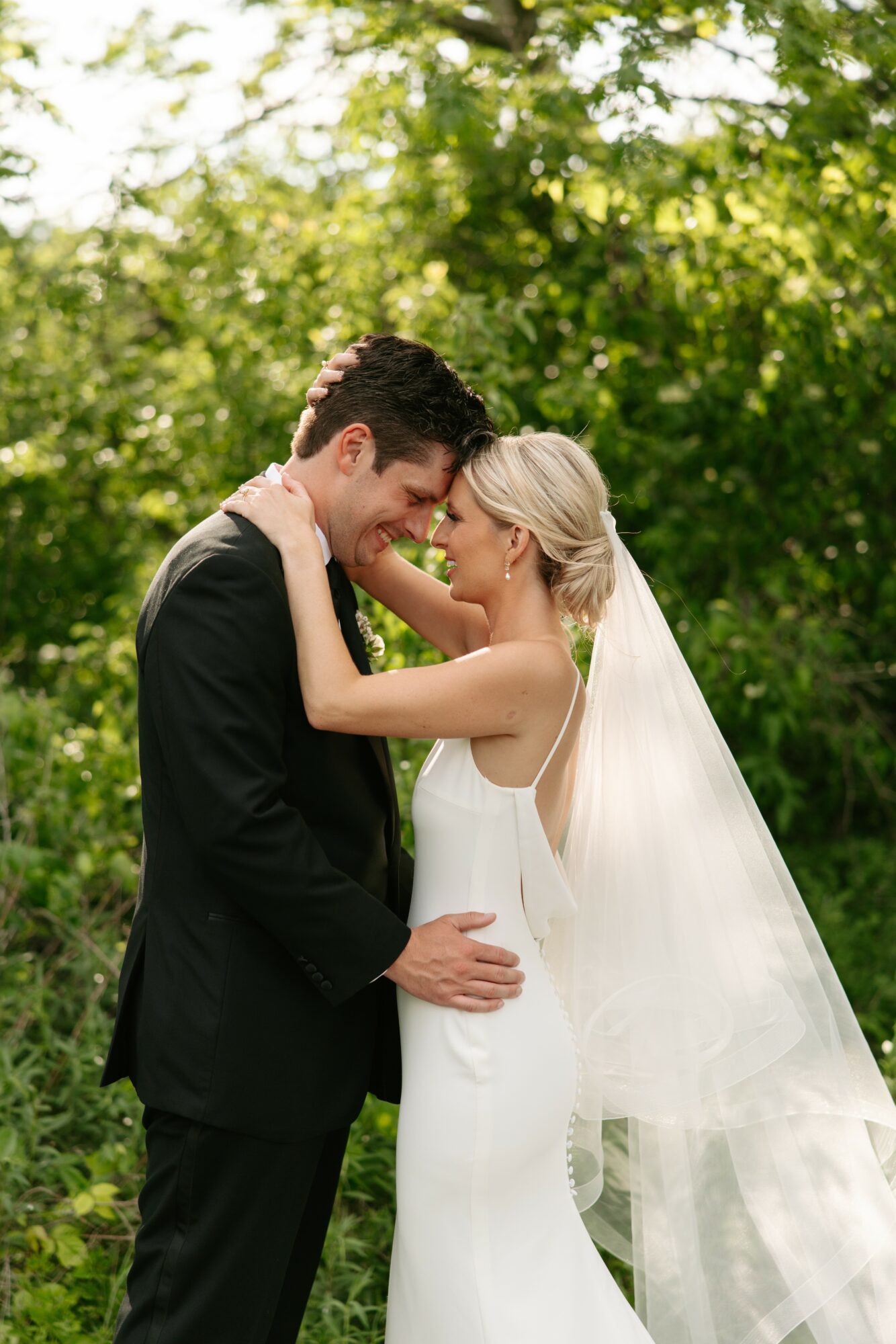 Bride and groom smiling and touching foreheads outdoors, surrounded by green trees, bride in white dress with veil, groom in black suit.