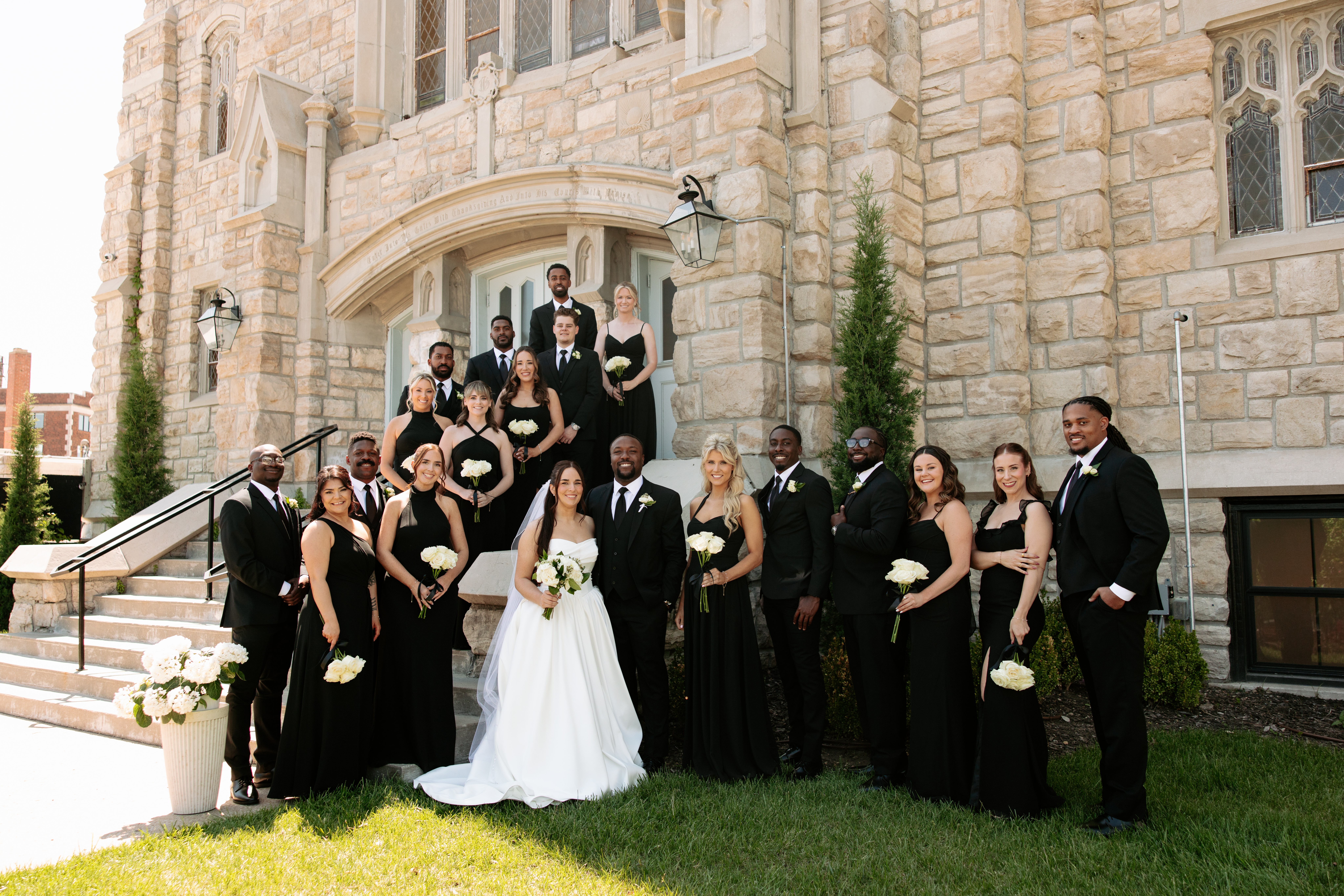 Group of people in formal attire standing outside a stone building, with some on stairs and others on the grass, celebrating a wedding.